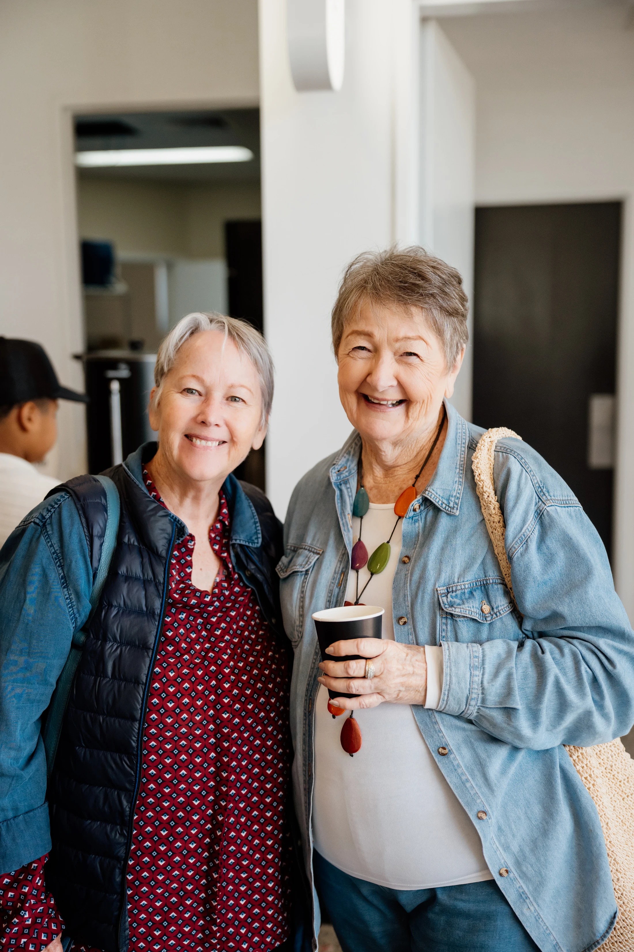 Two older women with short gray hair stand close together and smile for the camera. The woman on the right holds a black paper cup and wears a denim jacket.