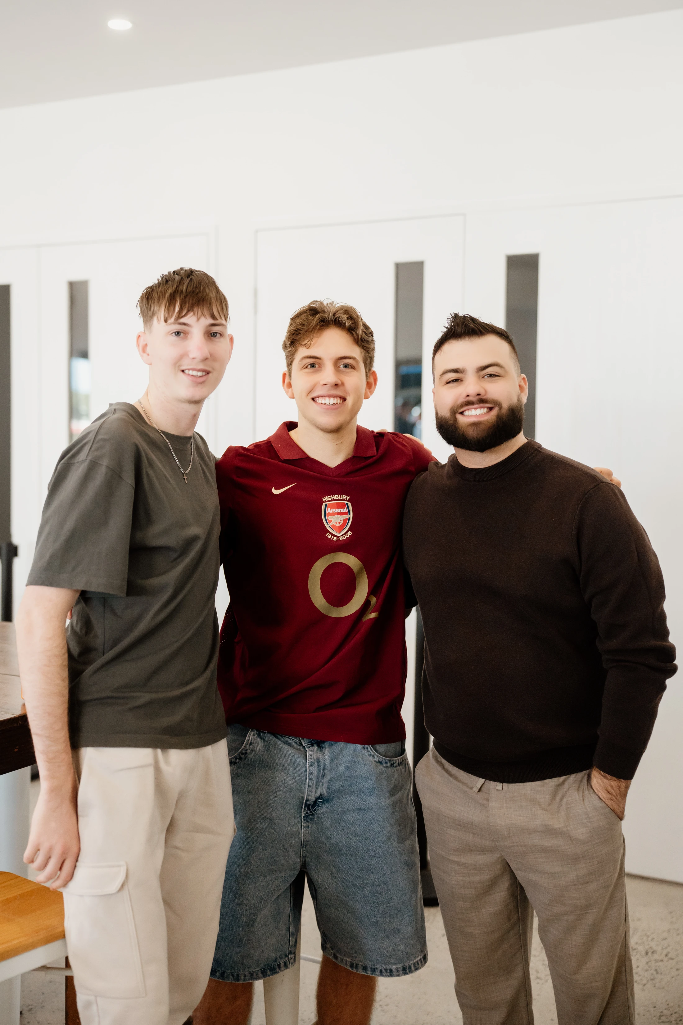 Three young men stand close together indoors, smiling at the camera. They have their arms around each other in a friendly pose.