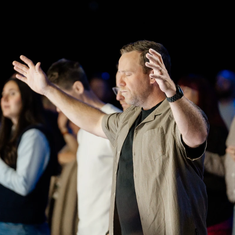 A man with his eyes closed and hands raised in a posture of worship stands in a crowd of people in a dimly lit room.