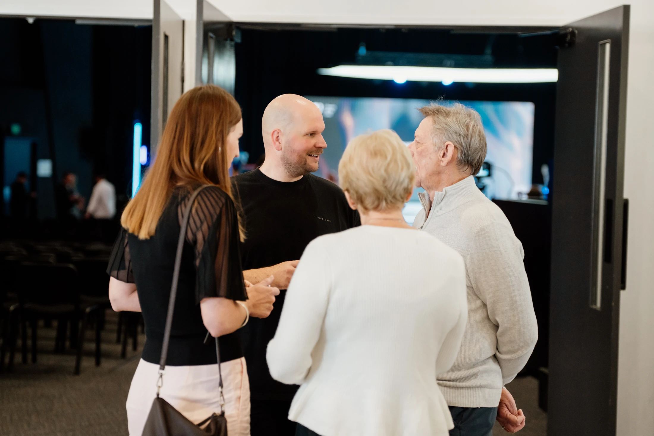 A group of four people, two men and two women of varying ages, stand together in a circle having a pleasant conversation in a modern lobby.