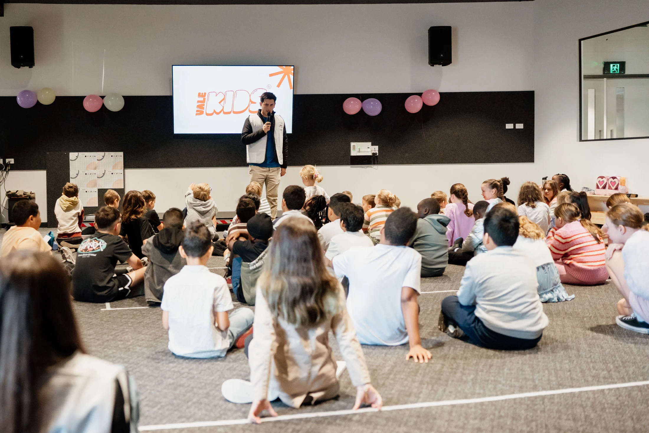 A man with a microphone speaks to a large group of children sitting on a gray carpet. Behind him is a screen with the 'Vale Kids' logo.