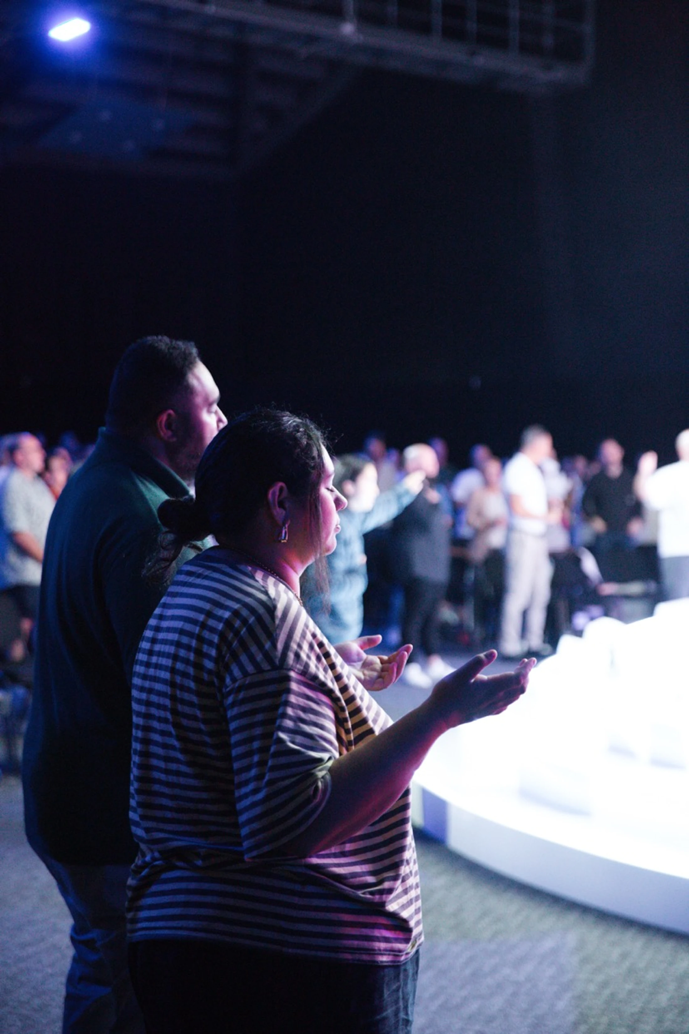 A woman in a striped shirt stands with her hands raised in worship among a crowd in a dimly lit room with purple and blue lighting.