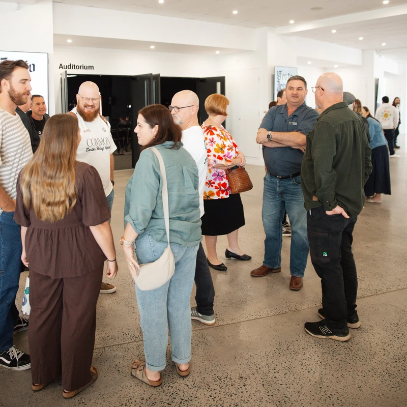 Groups of casually dressed adults stand and talk in a bright, modern building lobby with a sign for an auditorium in the background.