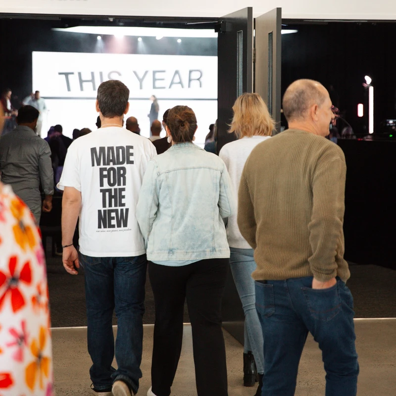 A view from behind of people walking into an event. One man wears a white shirt that reads 'Made For The New.' A screen in the background says 'This Year.'