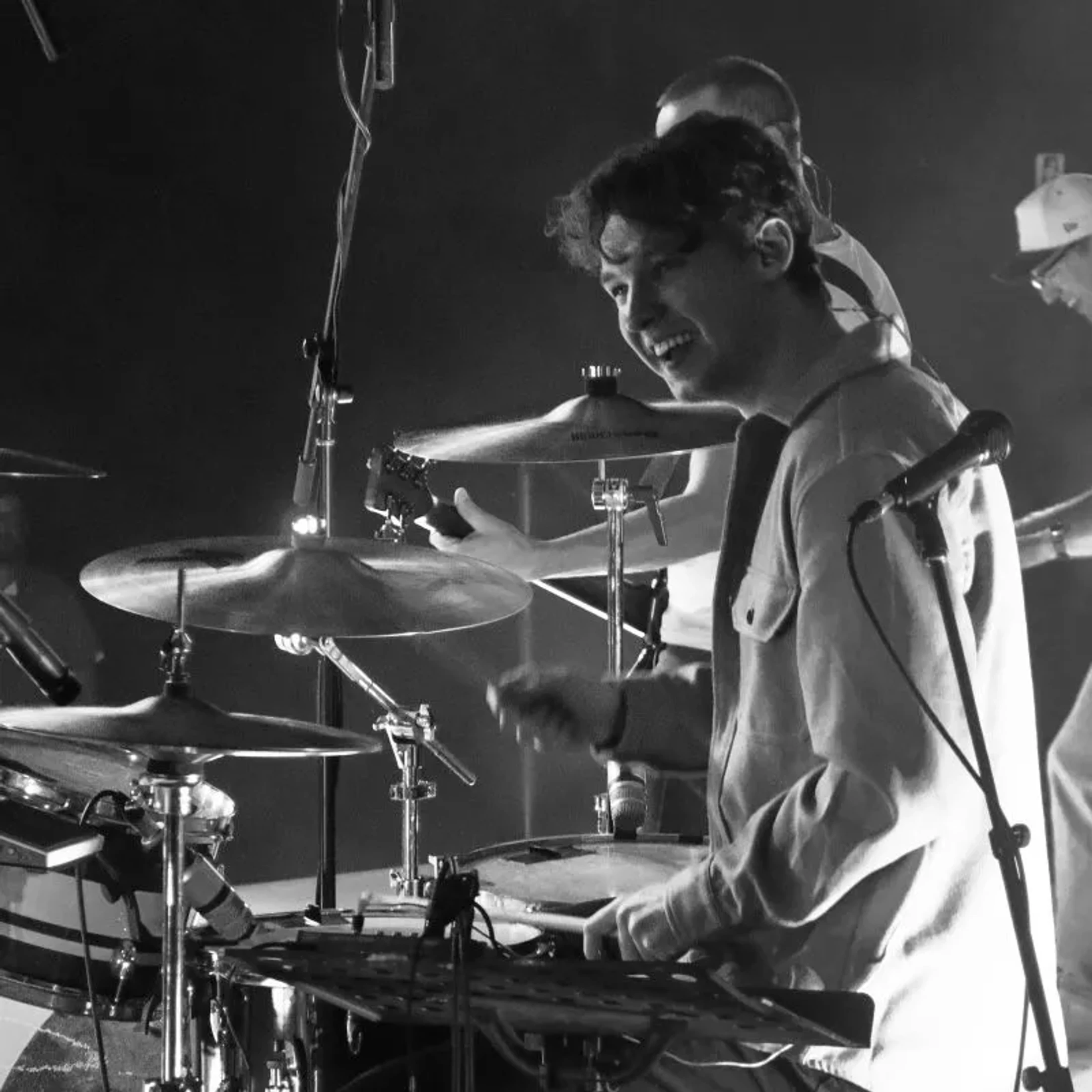 A young man smiles as he plays the drums on stage. The photo is in black and white, capturing a moment from a live musical performance.