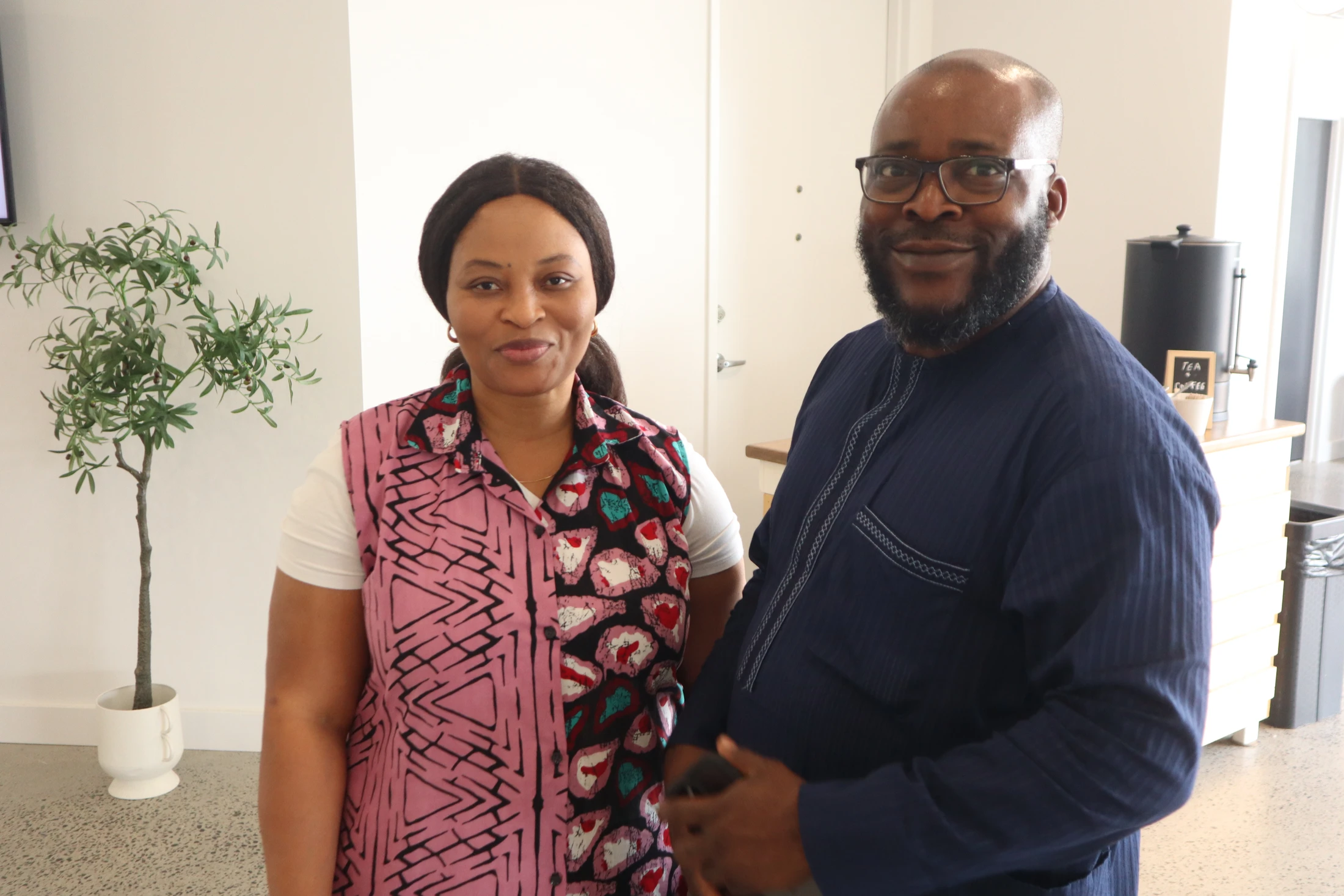 A man and a woman stand next to each other smiling for the camera in a modern room. The woman wears a colorful vest and the man wears a traditional blue shirt.