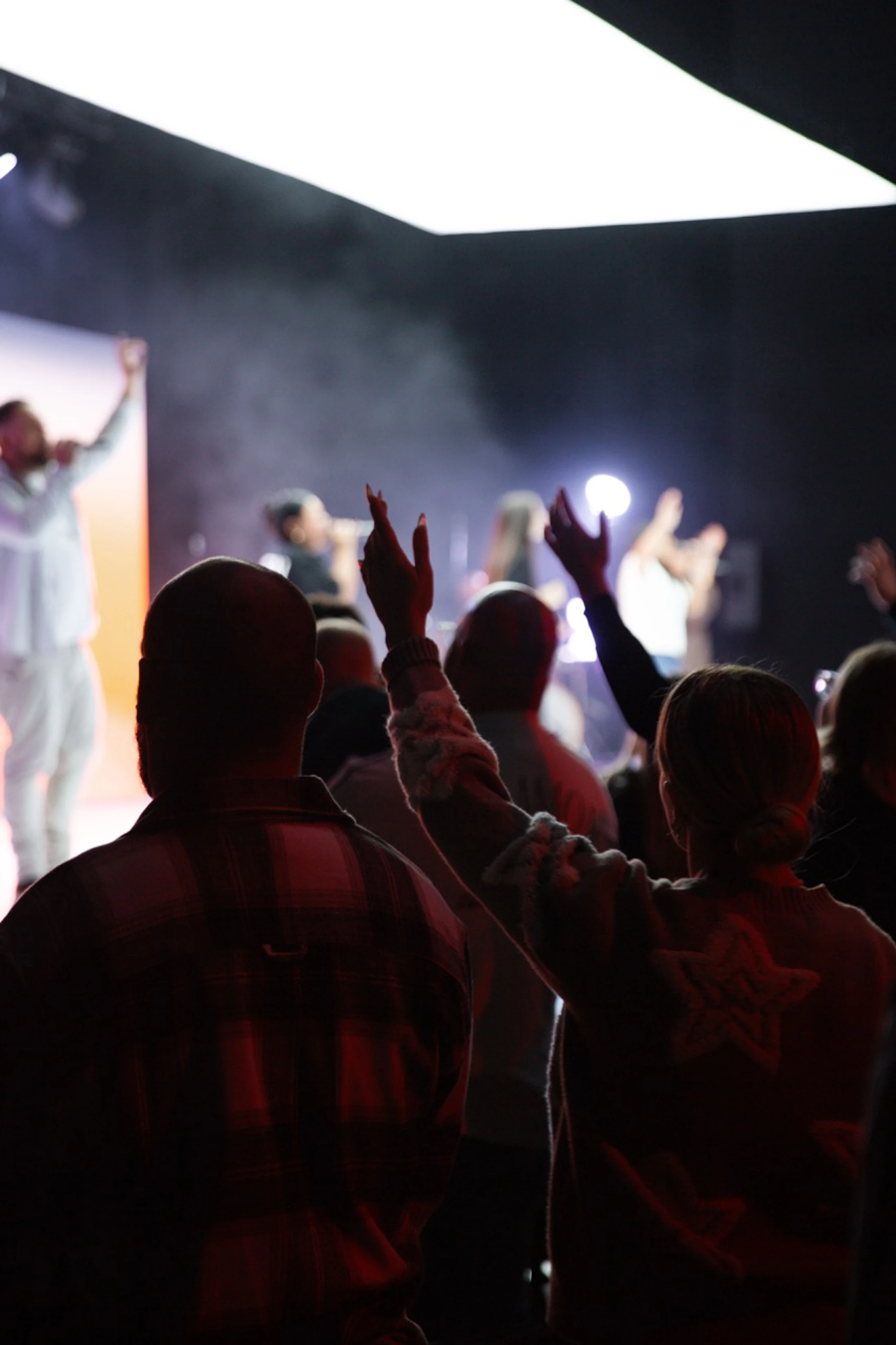 View from behind a crowd of people raising their hands in worship towards a brightly lit stage with a band and atmospheric smoke.
