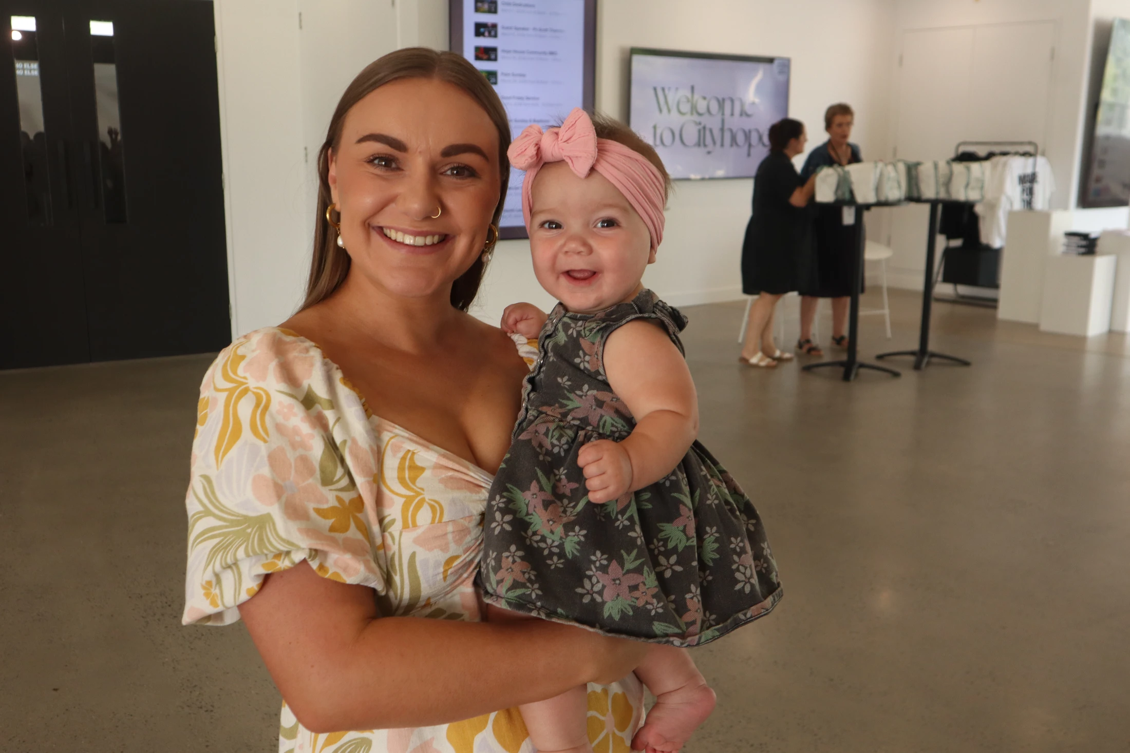 A woman in a floral dress smiles while holding a baby girl in a dark floral dress and a pink bow headband. They are indoors at an event.