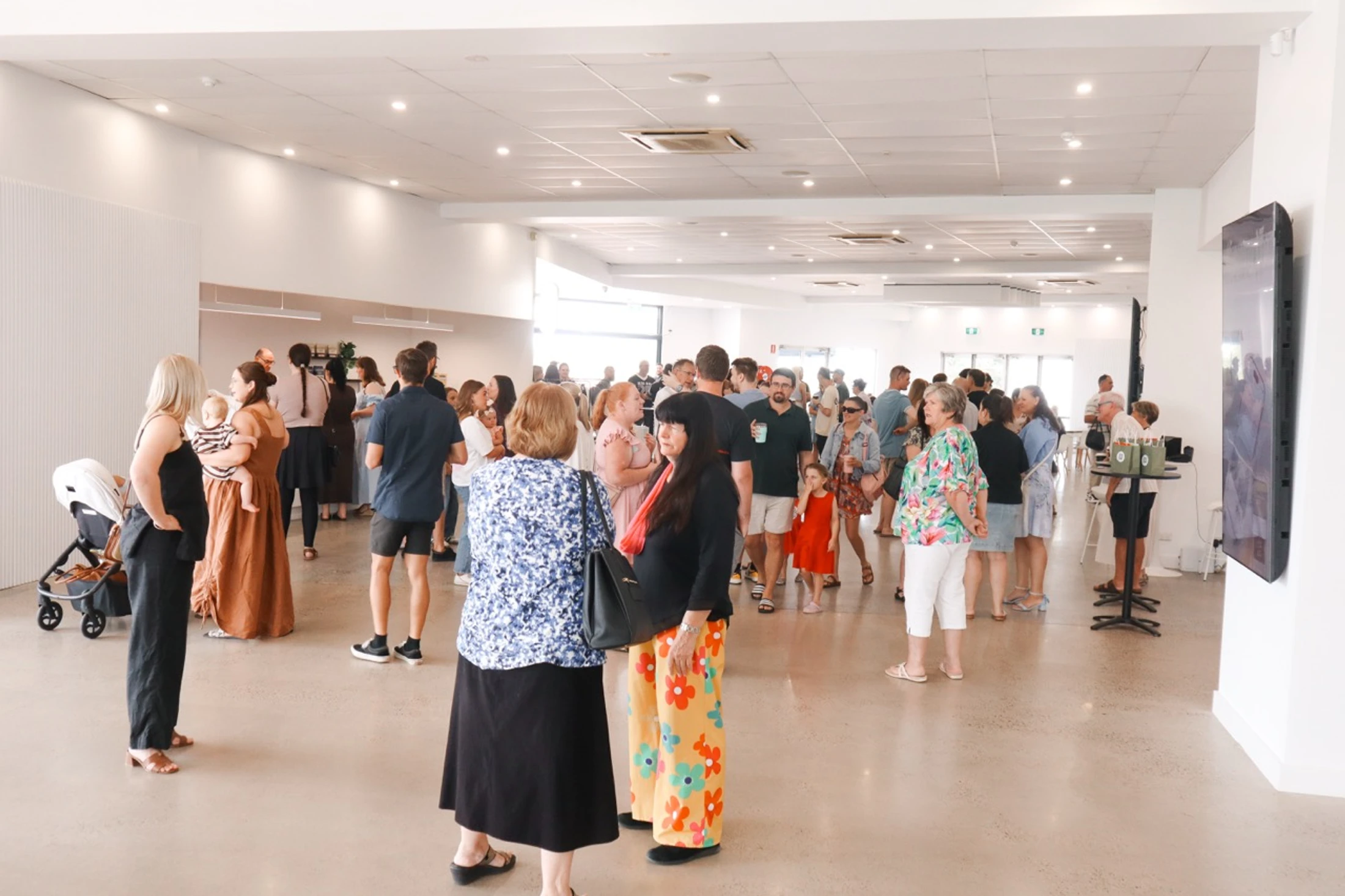 A wide shot of a brightly lit, modern hall filled with a diverse group of people standing and talking. They are dressed casually and appear to be socializing.