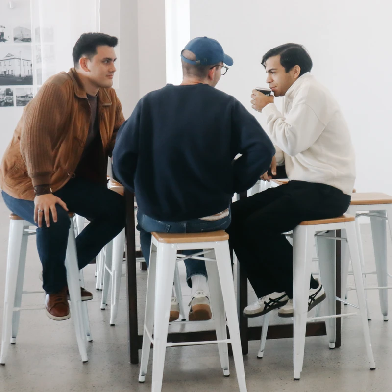 Three young men in casual clothes sit on tall white stools around a small table, engaged in conversation. One man drinks from a paper cup.
