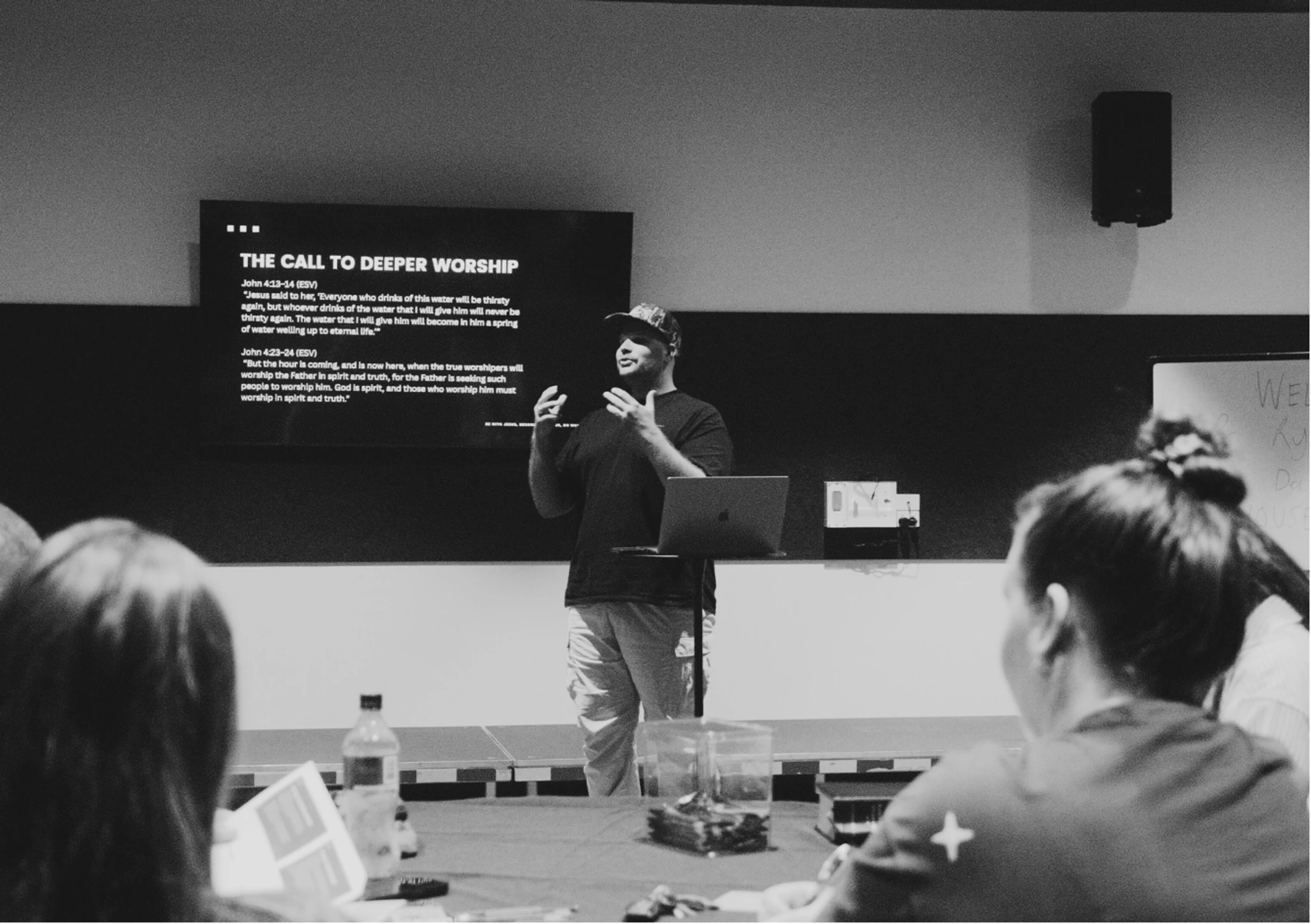 A black and white photo of a man in a cap teaching a class. A screen behind him reads 'THE CALL TO DEEPER WORSHIP' with Bible verses below.