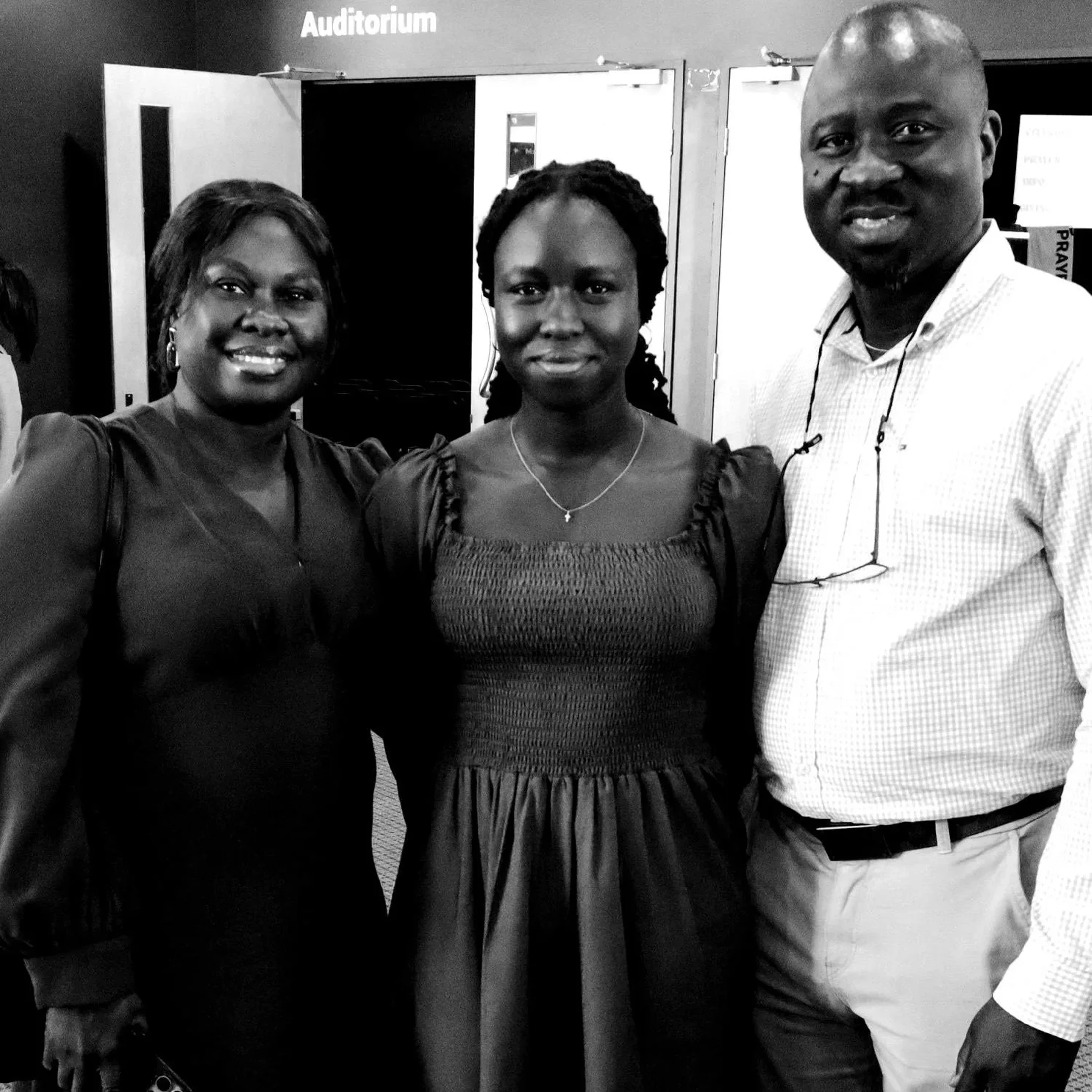 A black and white portrait of a smiling family of three standing together in front of a doorway labeled 'Auditorium'.