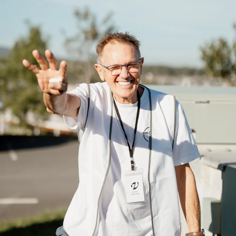 An older man with glasses smiles warmly while waving at the camera. He is outdoors on a sunny day, wearing a white volunteer vest and a name tag.