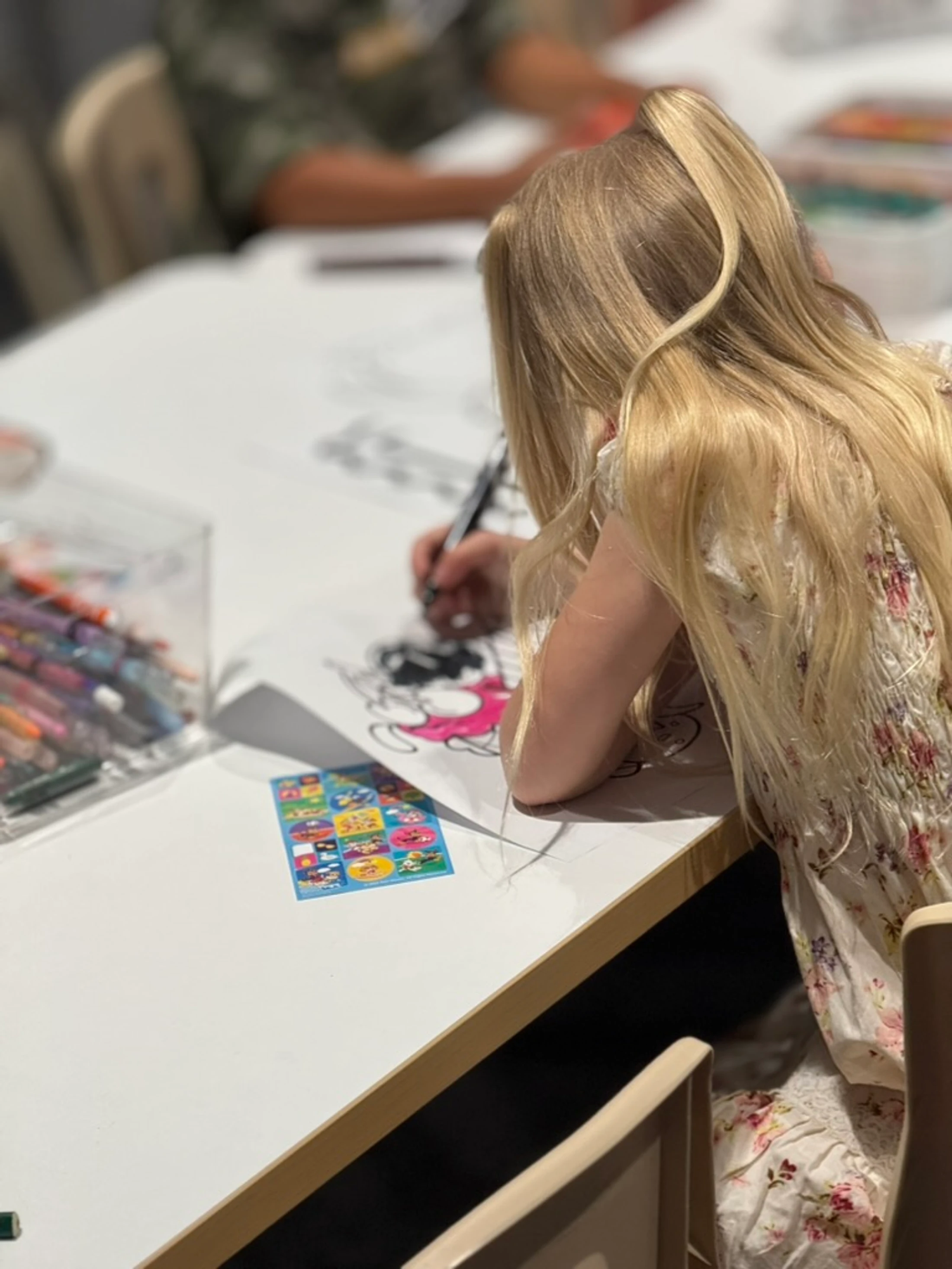 A girl with long blonde hair in a high ponytail leans over a white table, concentrating on coloring in a book. A box of crayons is nearby.