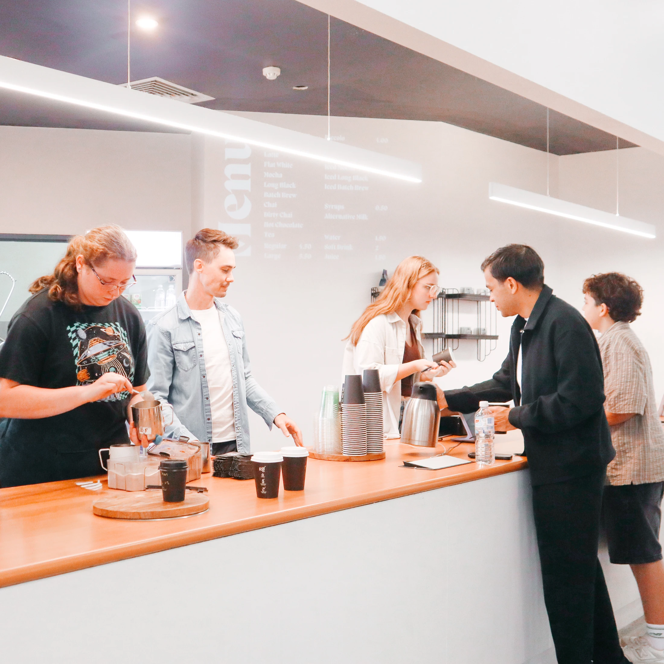 A brightly lit coffee shop with a barista making drinks and several customers ordering and waiting at a long wooden counter.