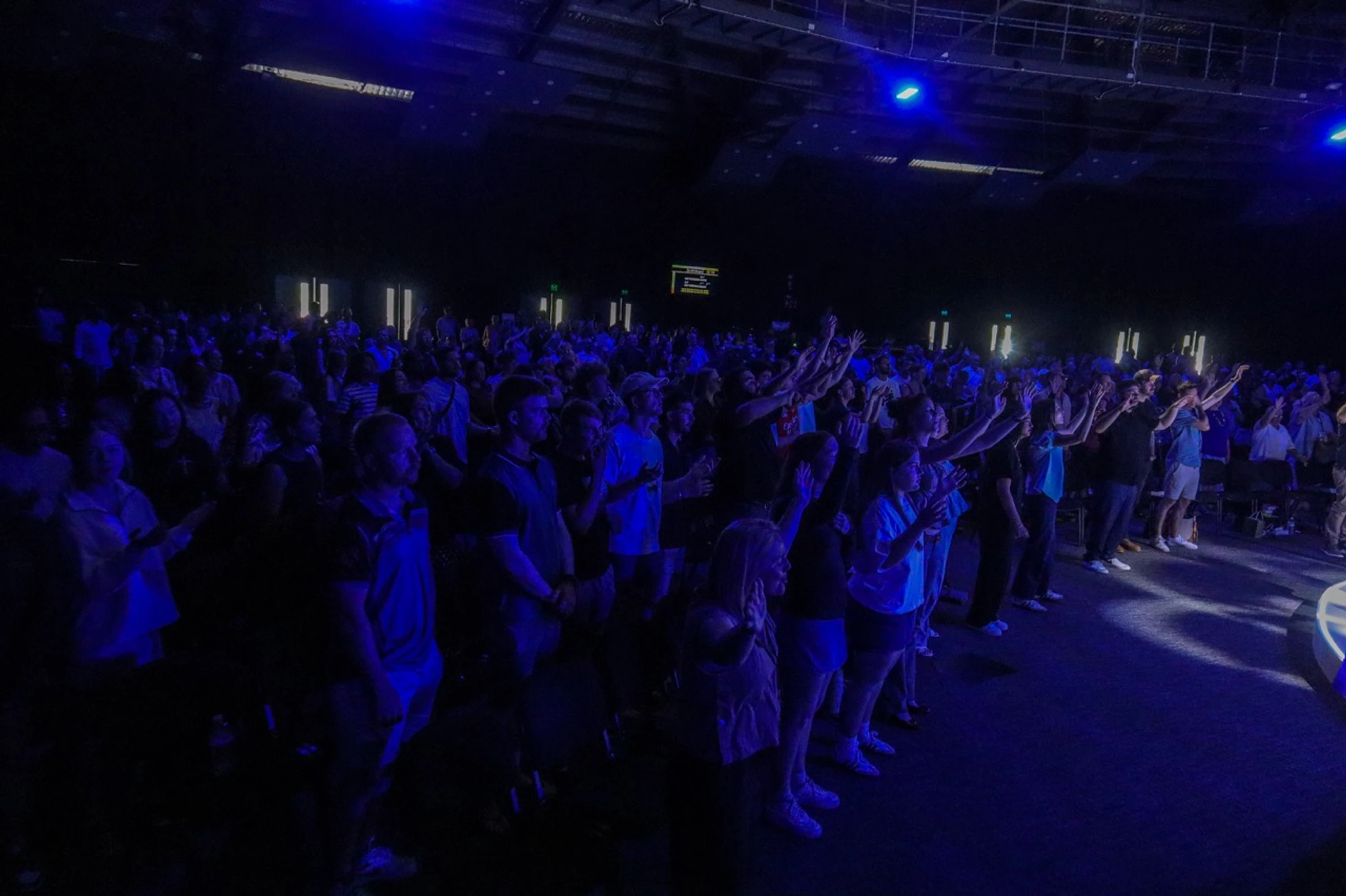A large crowd of people stands in a dark auditorium, illuminated by blue stage lights. Many have their hands raised in an act of worship or praise.
