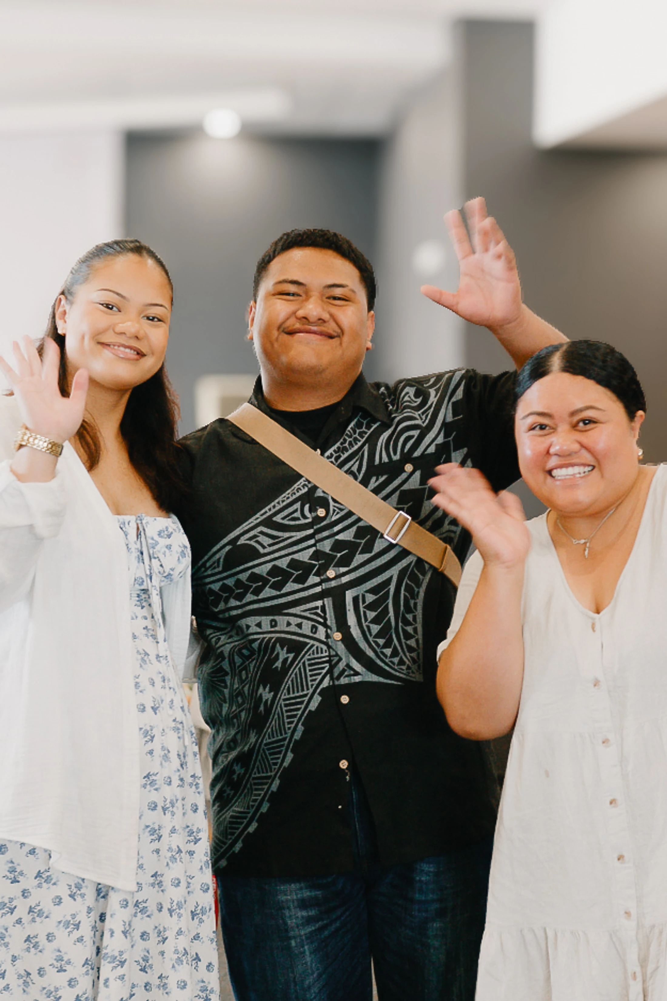 A man stands between two women as they all smile and wave at the camera. The man wears a shirt with a Polynesian pattern.