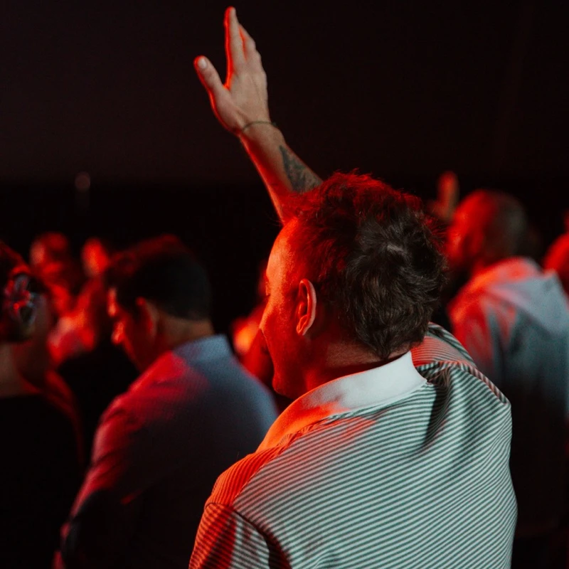 A man seen from behind in a dark, red-lit room raises his tattooed arm. He wears a striped polo shirt and is surrounded by an out-of-focus crowd.