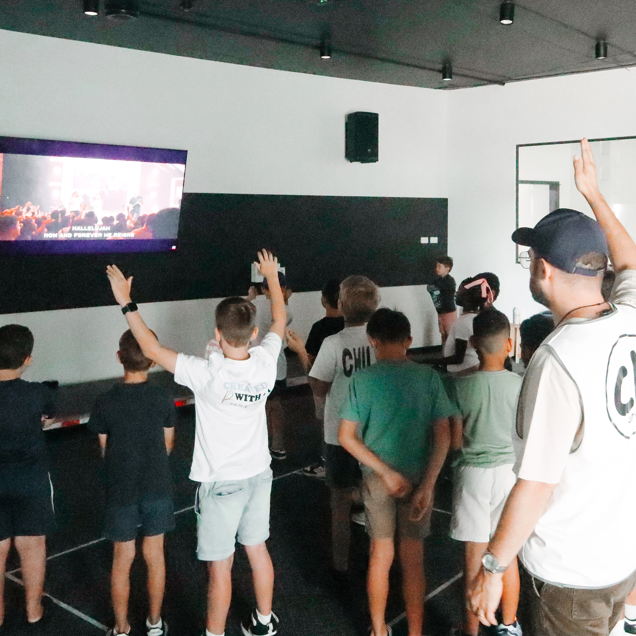 A group of children and an adult leader, seen from behind, worshiping with hands raised while watching a service on a large screen with lyrics.