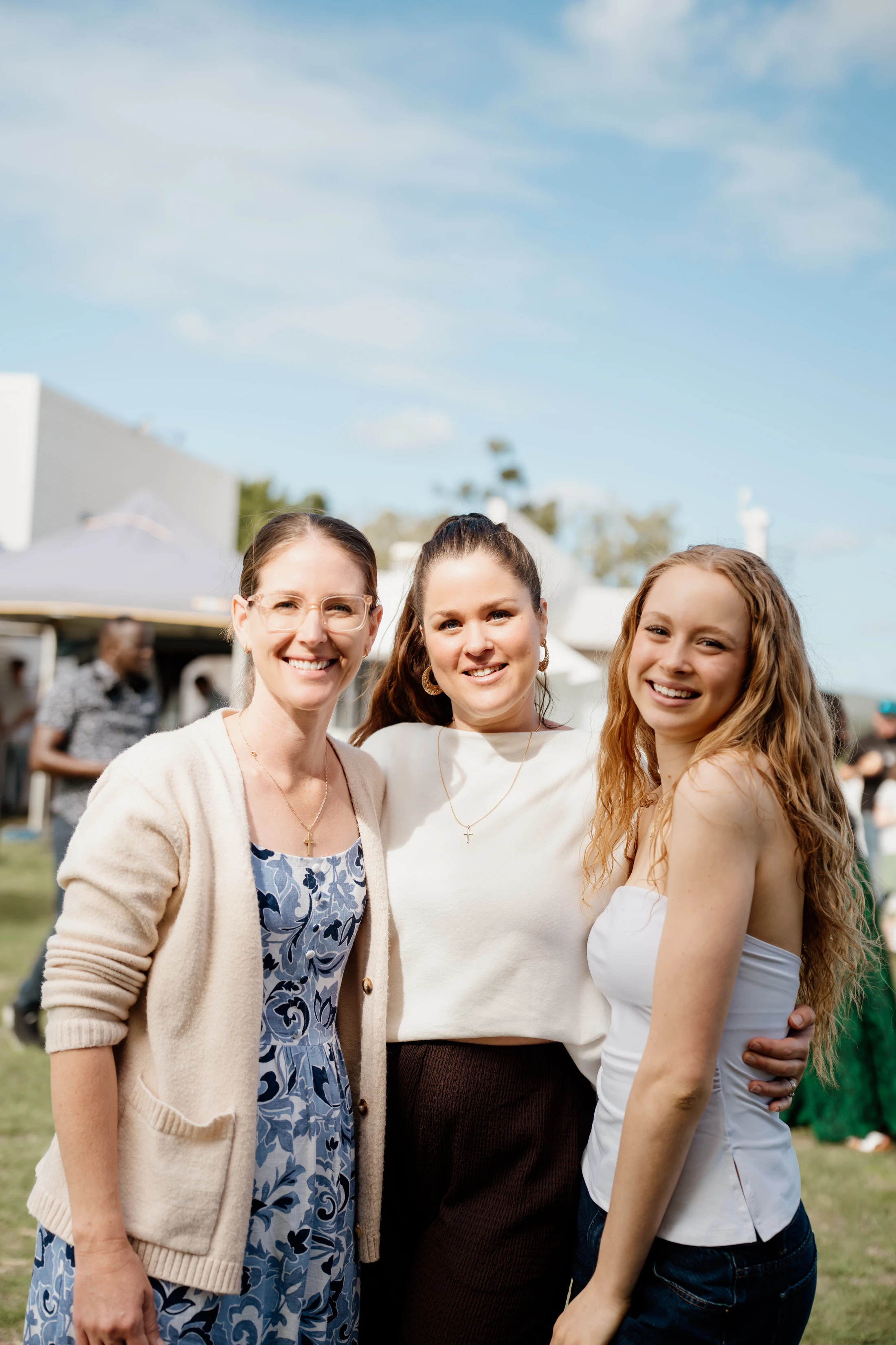 Three women stand close together, posing and smiling for a photo outdoors on a sunny day with a grassy field and blue sky in the background.