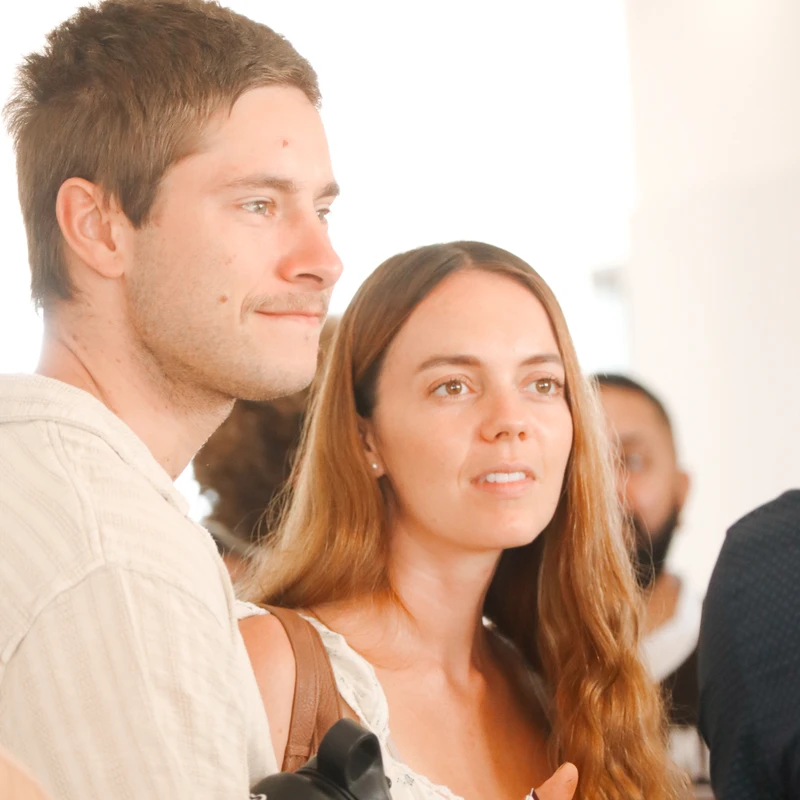 A young man and woman stand close together in a brightly lit indoor space, both looking off to the right with attentive expressions.