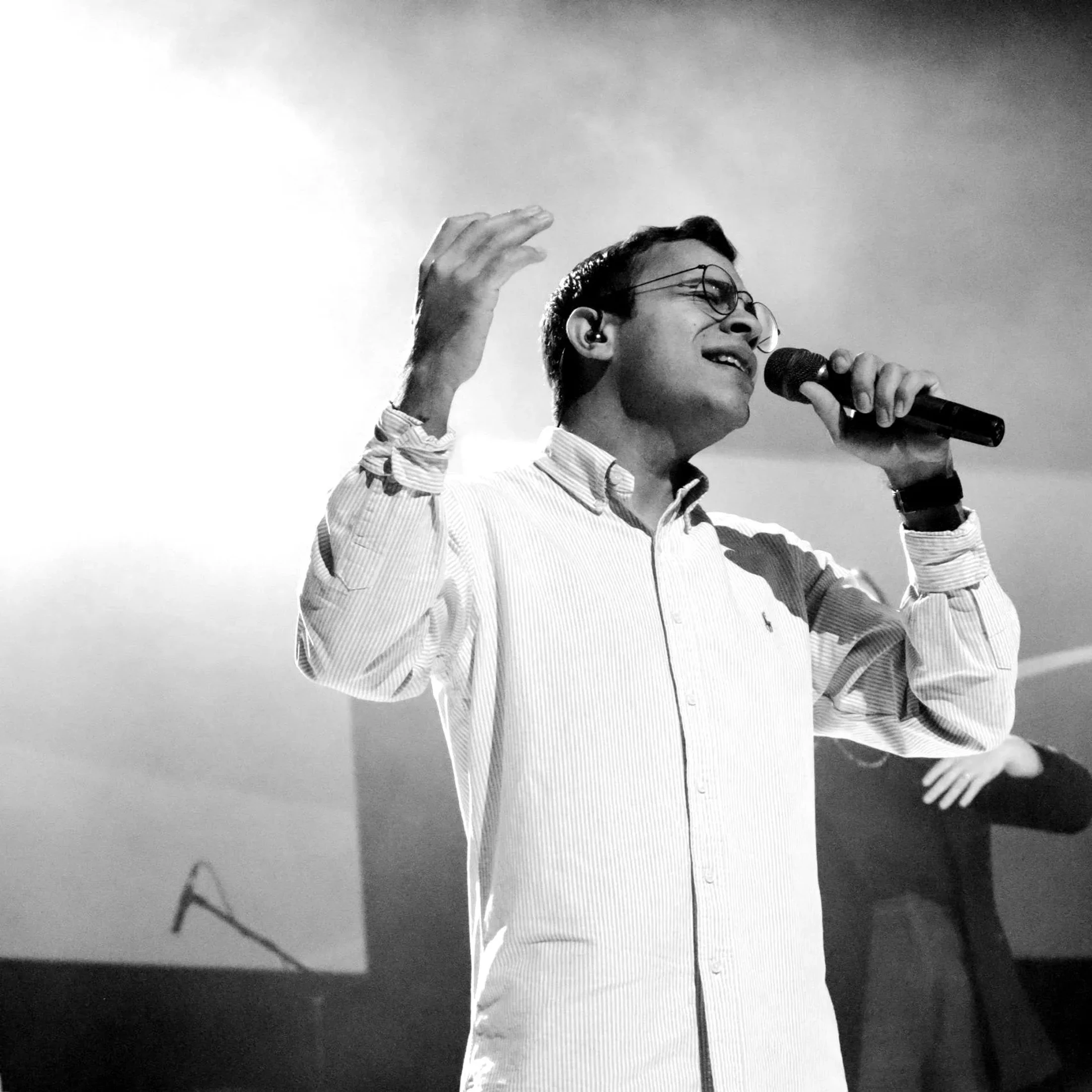 A black and white photo of a man in glasses and a striped shirt singing into a microphone on stage. He has one hand raised in a gesture of expression.