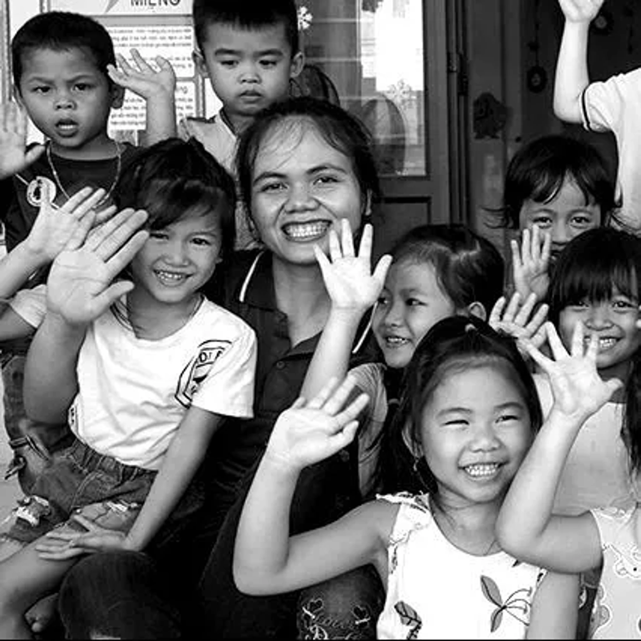 A black and white photo of a smiling woman surrounded by a group of happy children of various ages. They are all looking at the camera and waving.