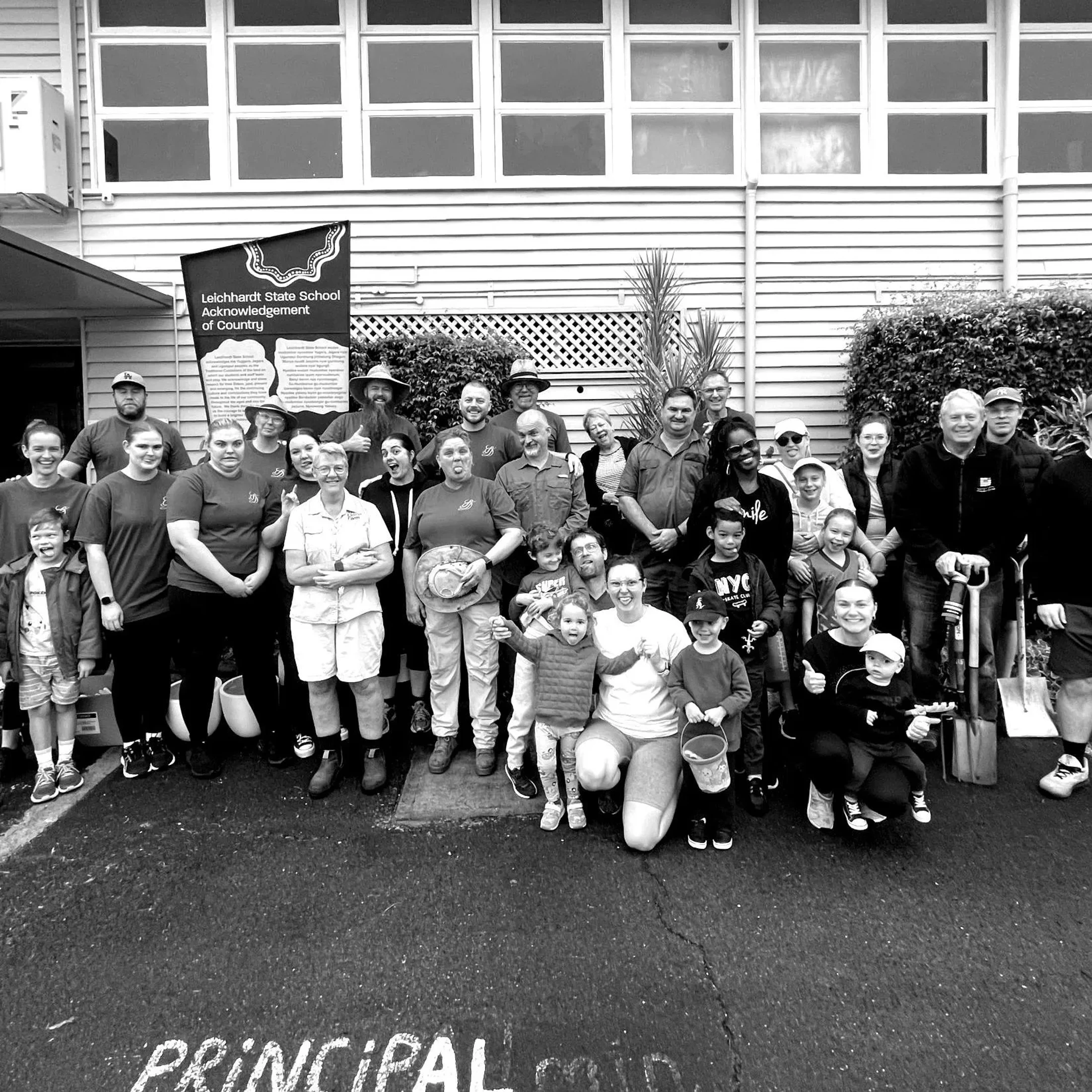 A large group of adults and children pose for a black and white photo in front of the Leichhardt State School building after a community working event.