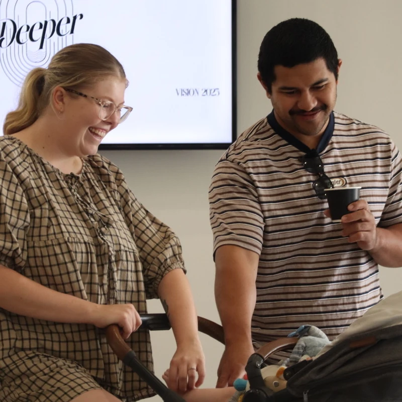 A woman in a plaid dress and a man in a striped shirt smile as they look down at a baby in a stroller. A screen in the background says "Deeper Vision 2025".