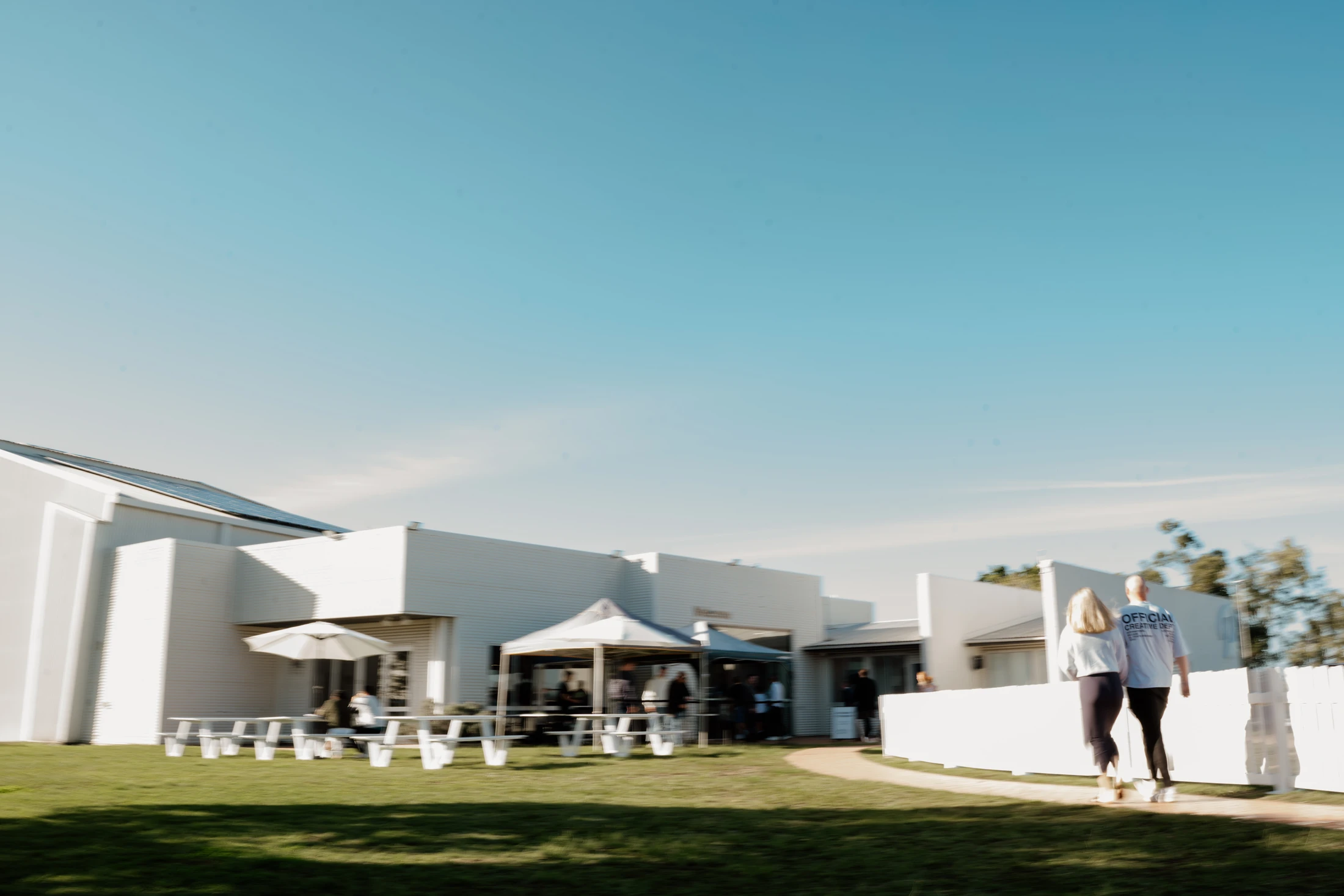 A motion-blurred photo of people at a modern white building with a lawn and picnic tables. A couple walks on a path under a clear blue sky on a sunny day.