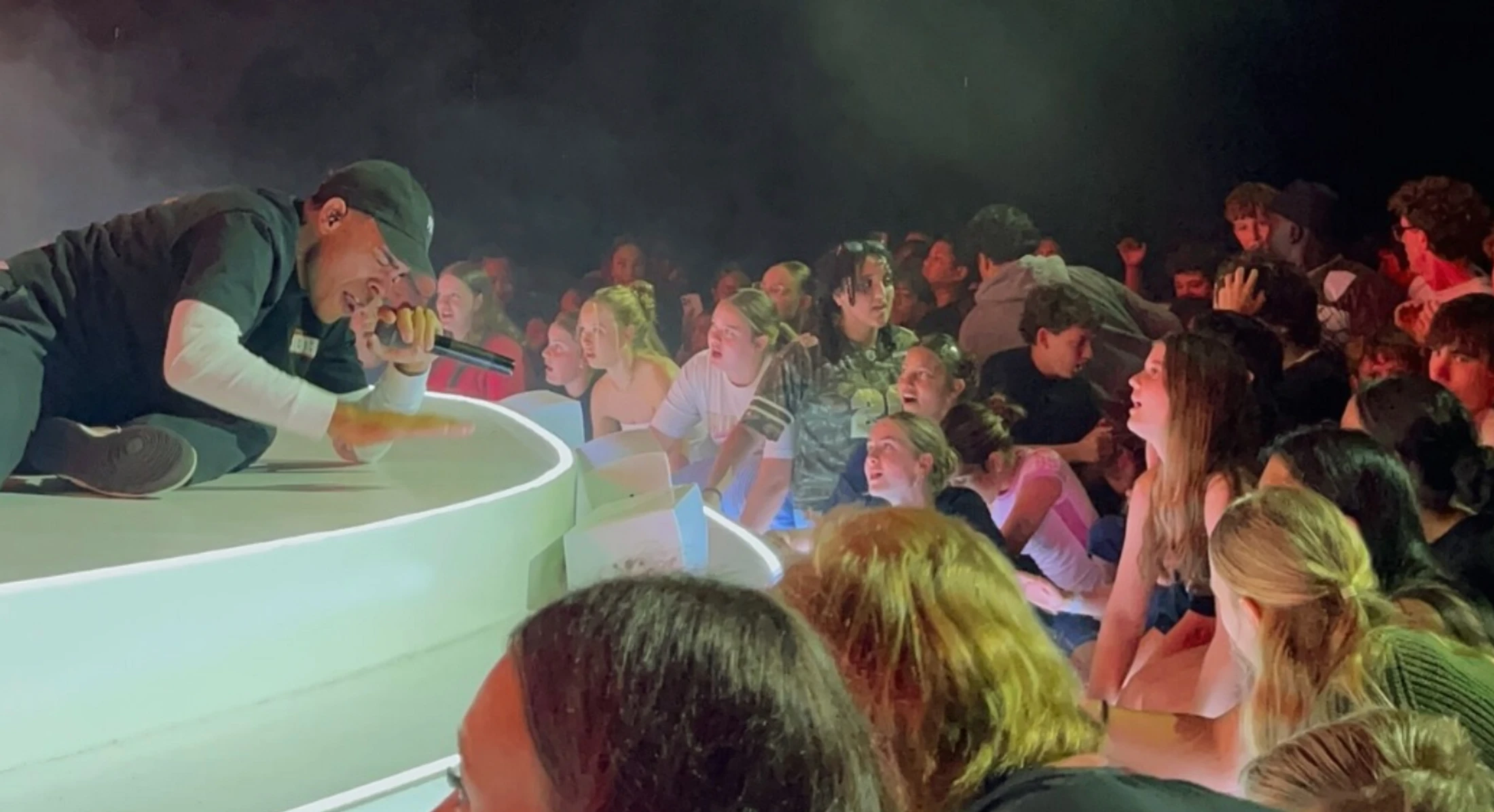A male rapper in a black cap kneels on a white stage, singing into a microphone inches from the faces of an excited young audience in the front row.