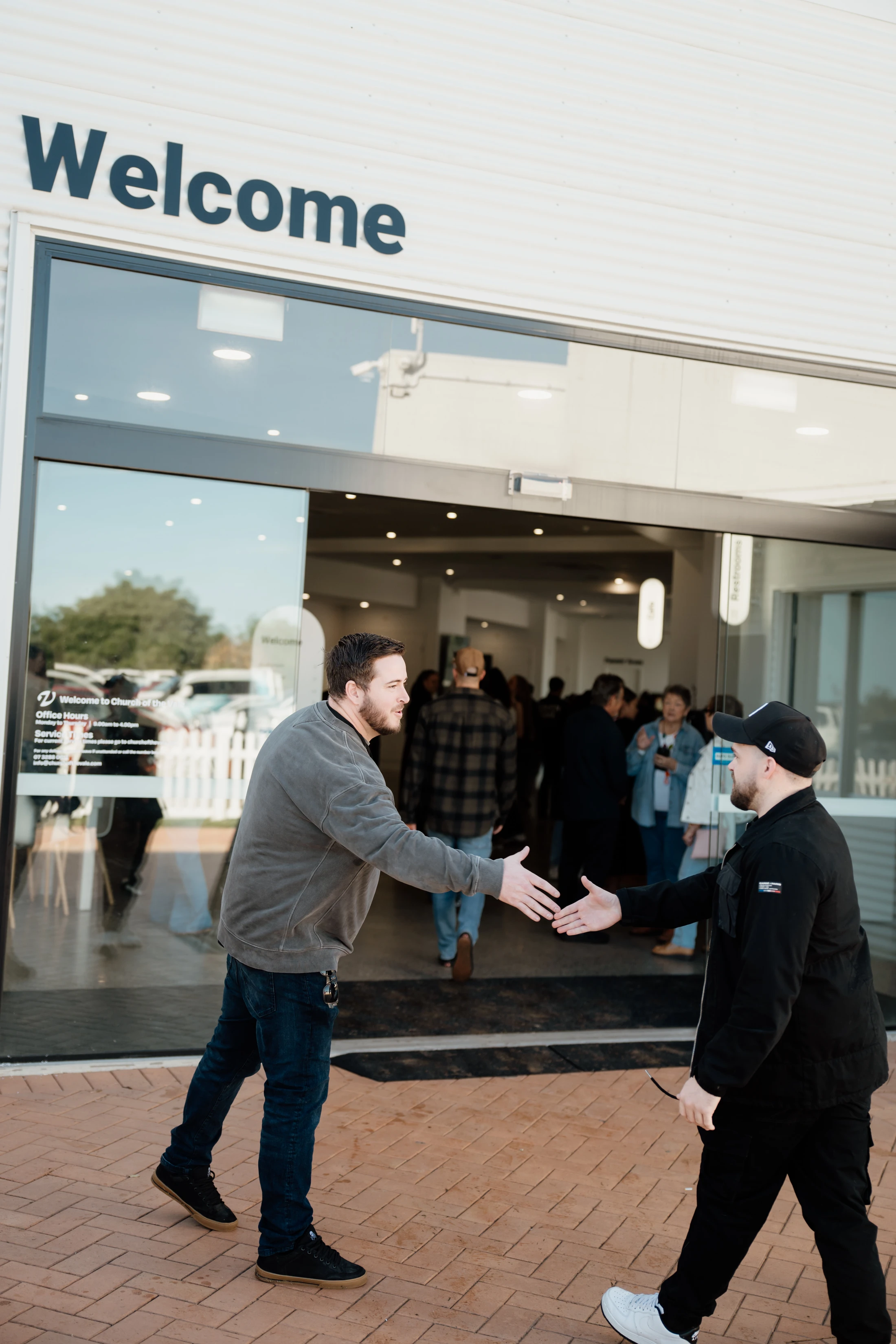 Two men shake hands outside the glass entrance of a modern building with a large 'Welcome' sign. Other people are visible inside and outside the doors.