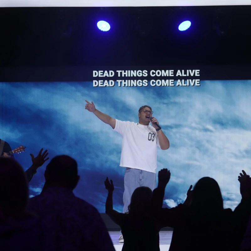 A man sings on stage in front of a screen with lyrics. In the foreground, silhouettes of the audience have their hands raised in worship.