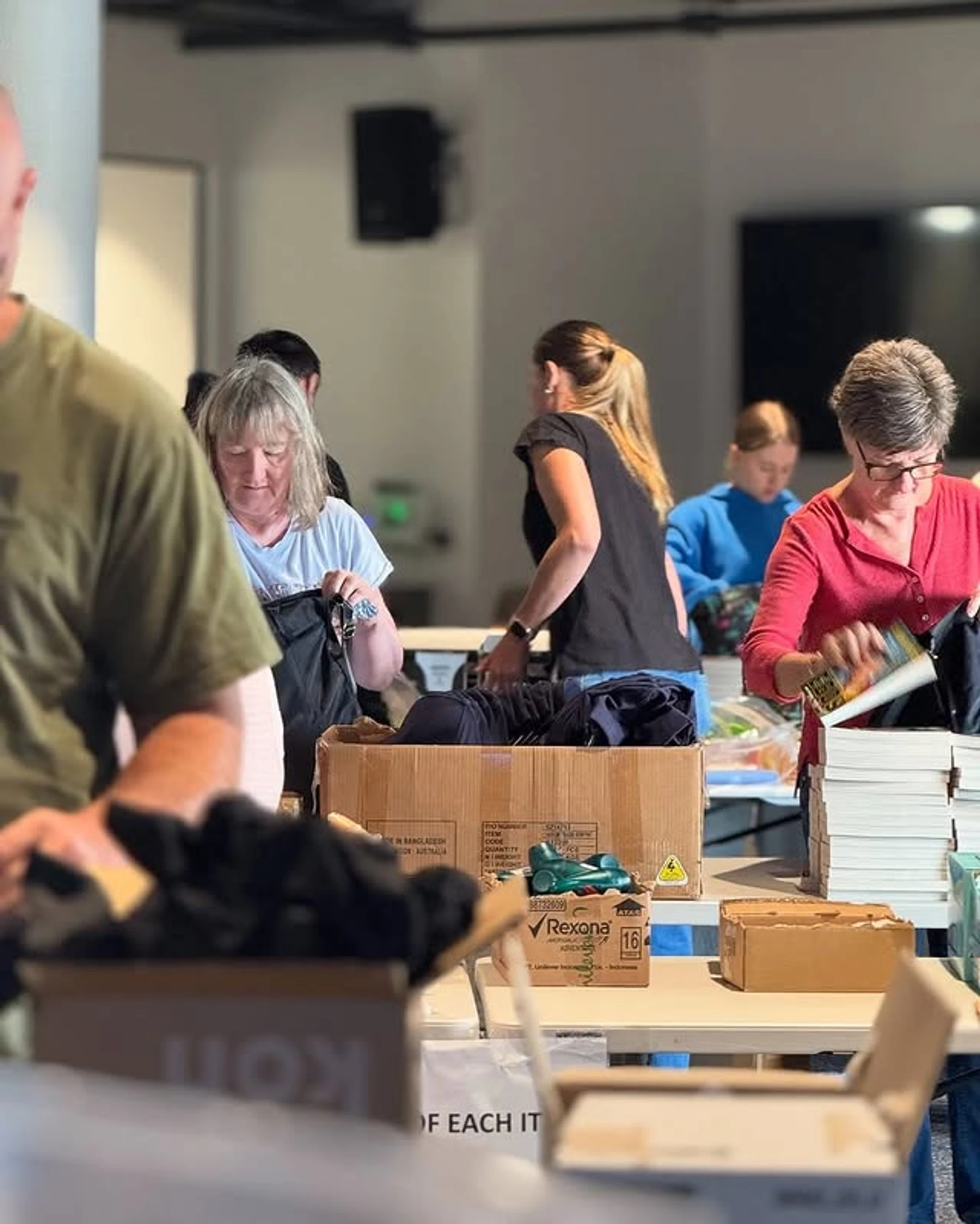 A group of people stand around tables indoors, sorting through donated items like books and clothes and packing them into large cardboard boxes.