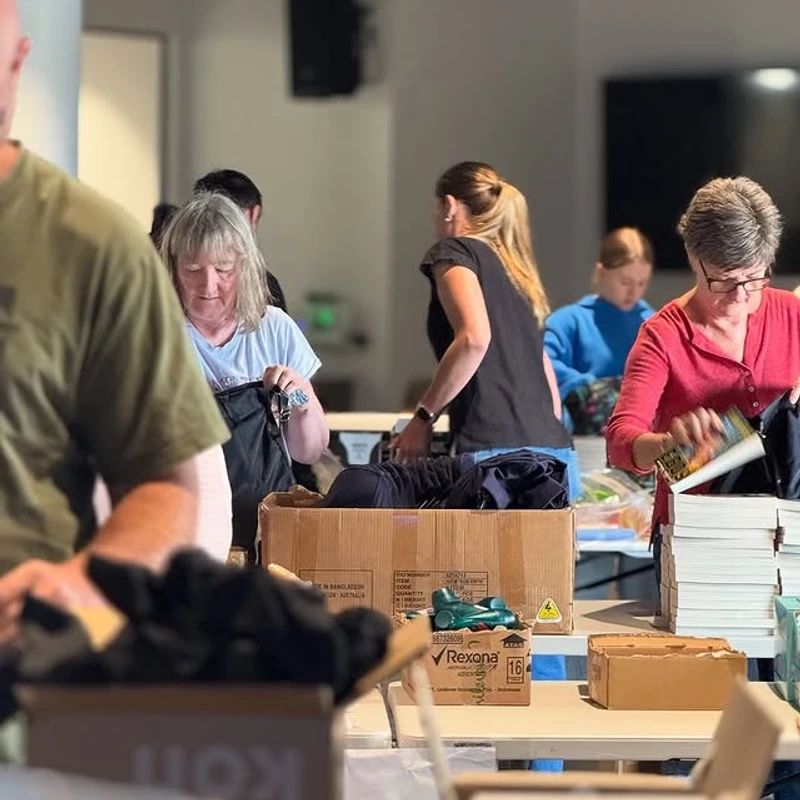 A group of people stand around tables indoors, sorting through donated items like books and clothes and packing them into large cardboard boxes.