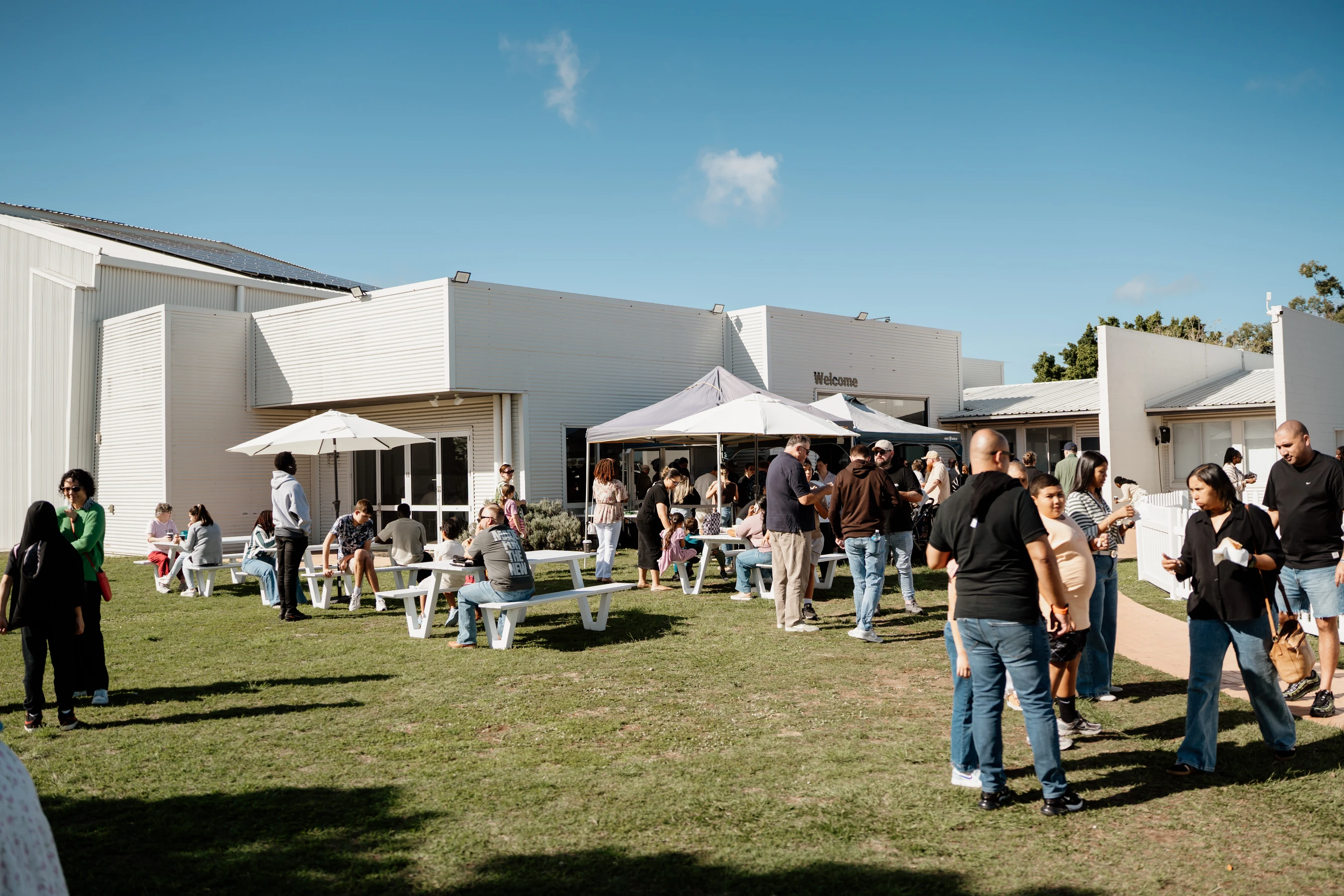 A diverse group of people gathered on a sunny day, standing and sitting on a lawn in front of a modern white building with a 'Welcome' sign.