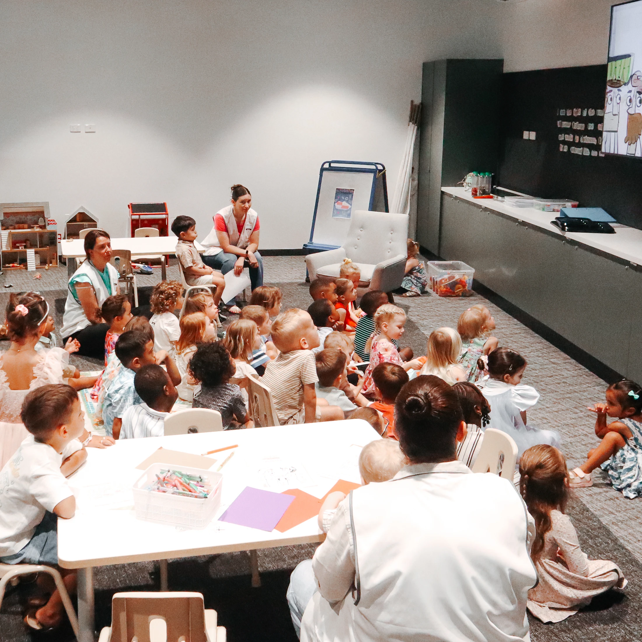A group of young children and toddlers sit on the floor and at small tables, watching a cartoon on a large screen with their teachers in a classroom.