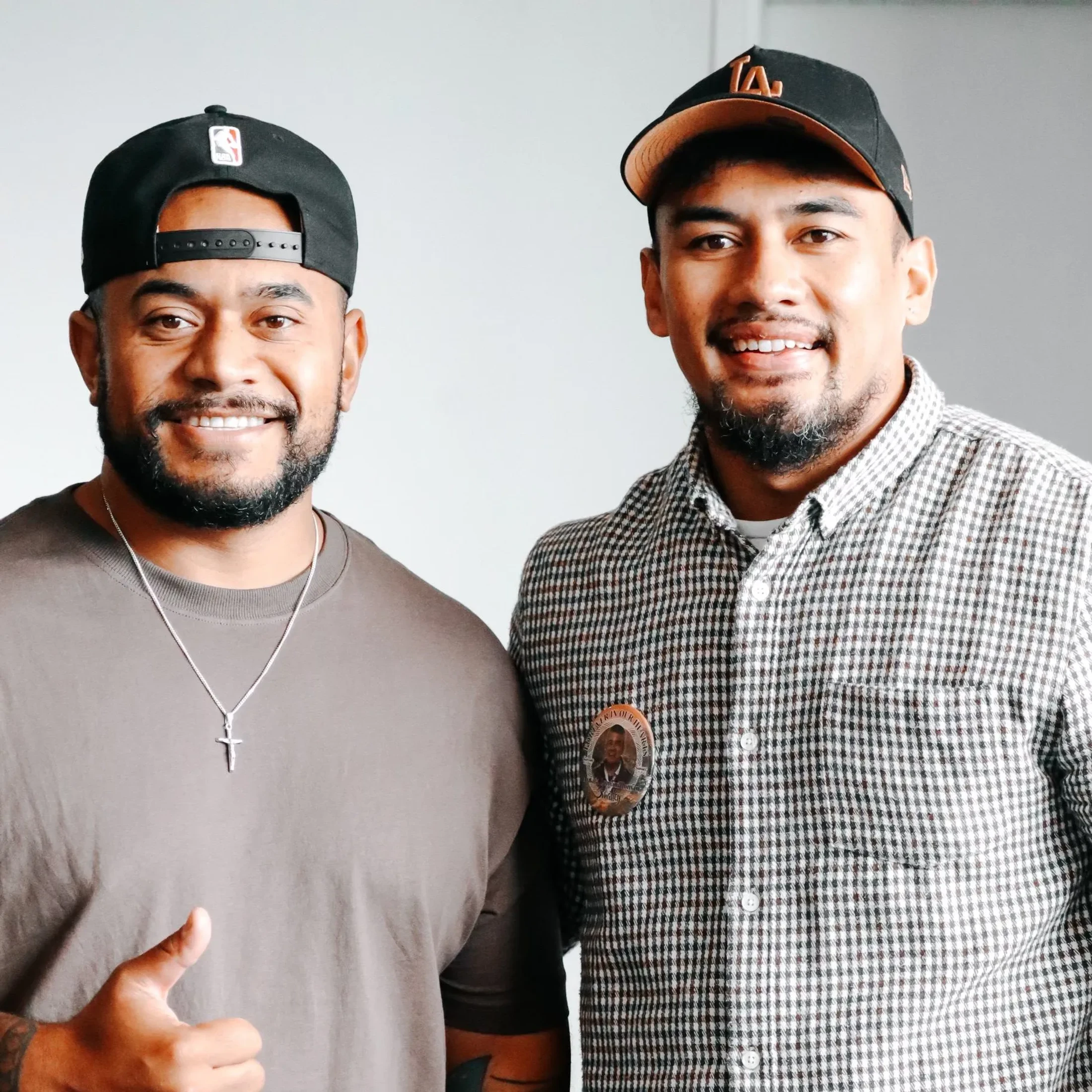 A head-and-shoulders portrait of two smiling men standing next to each other. Both have beards and are wearing baseball caps against a plain background.
