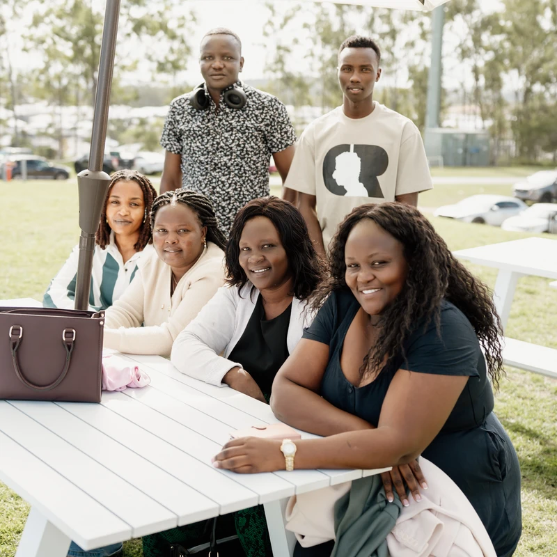 A group of six Black people smile for the camera while sitting and standing around a white picnic table with a large umbrella in a grassy park.