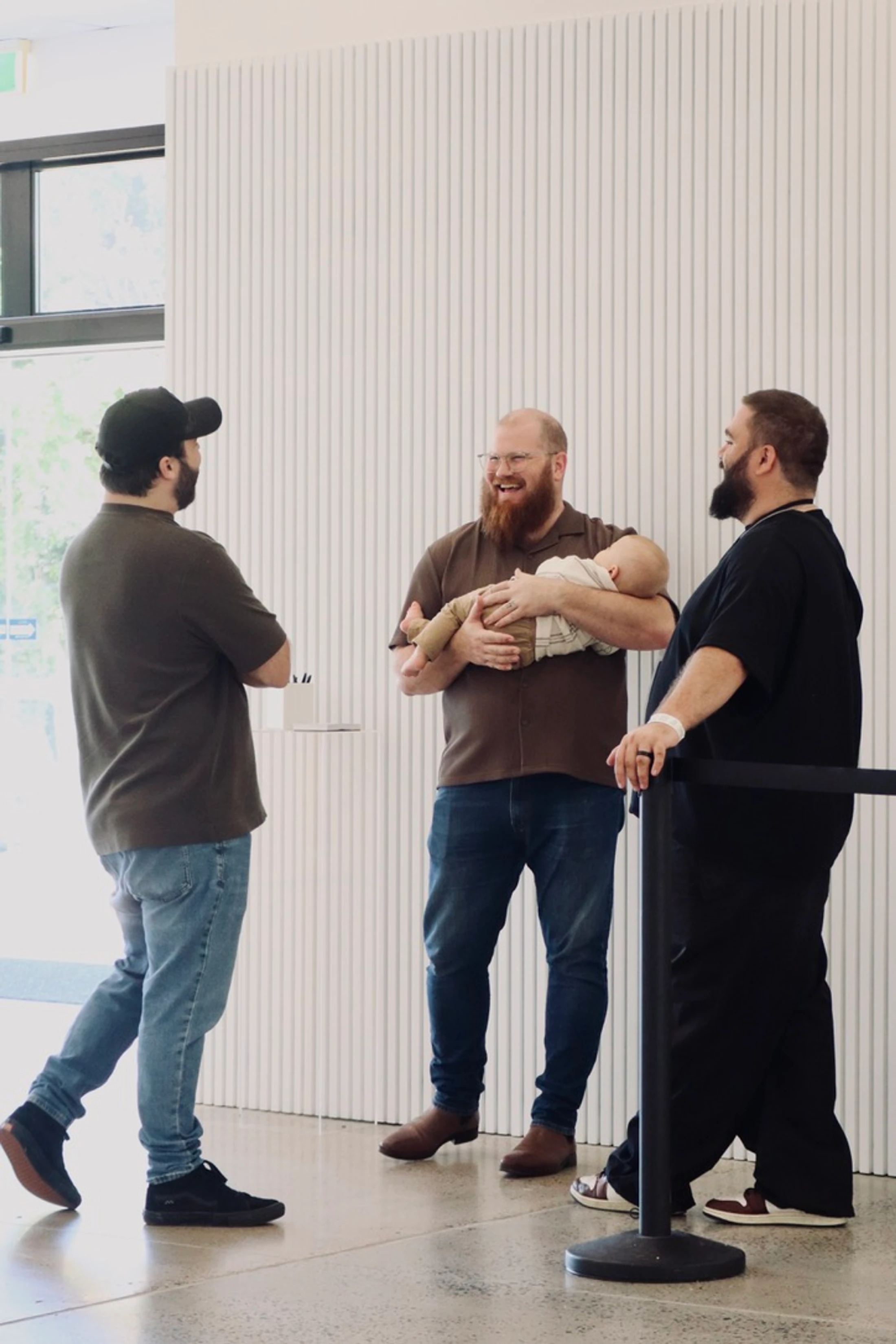 Three men stand talking in a brightly lit room. A smiling man with a red beard holds a sleeping baby, showing it to his two friends.