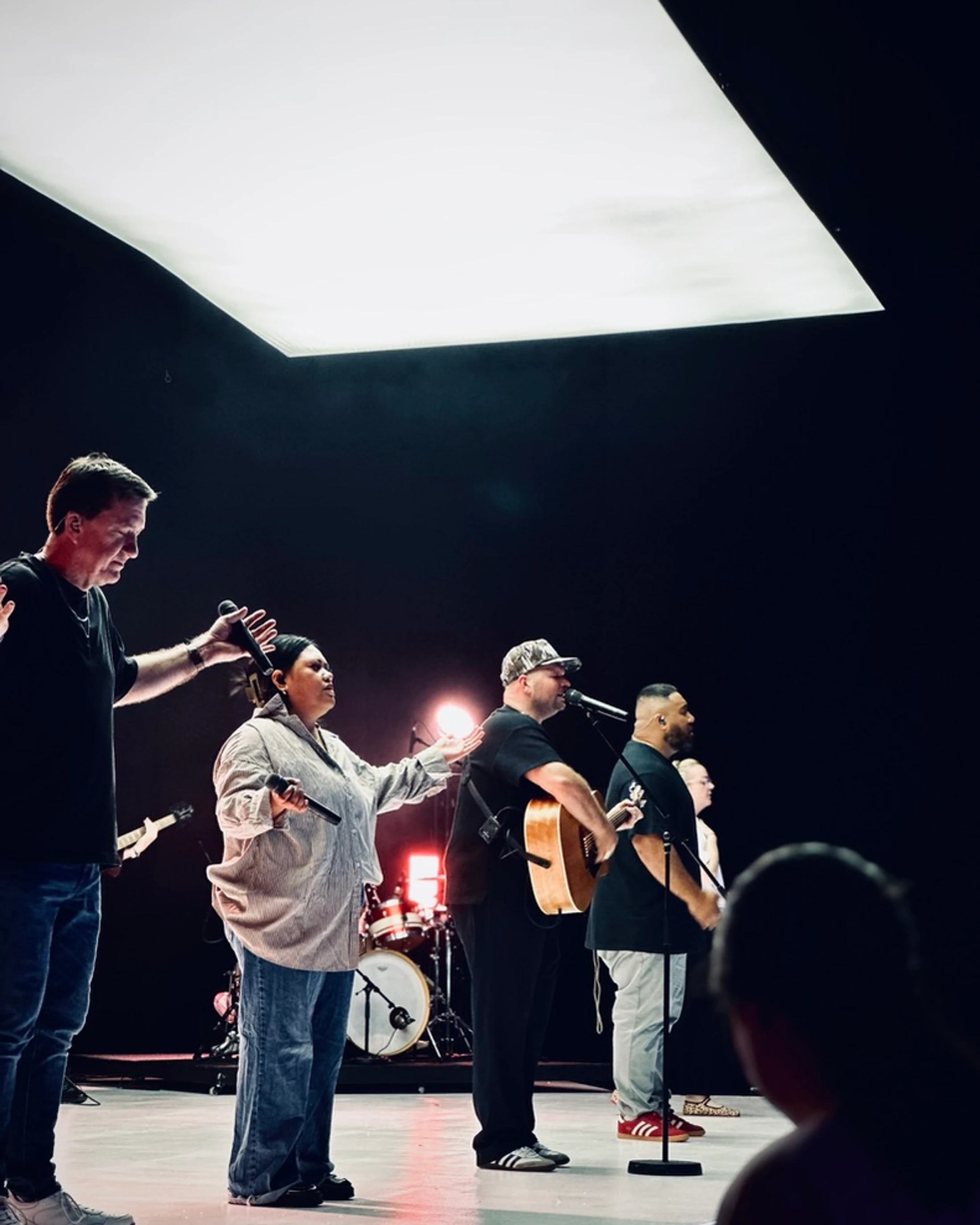 A diverse group of people stand on a dark stage under a large rectangular light, singing and playing music as a band, with one man placing a hand on a woman.