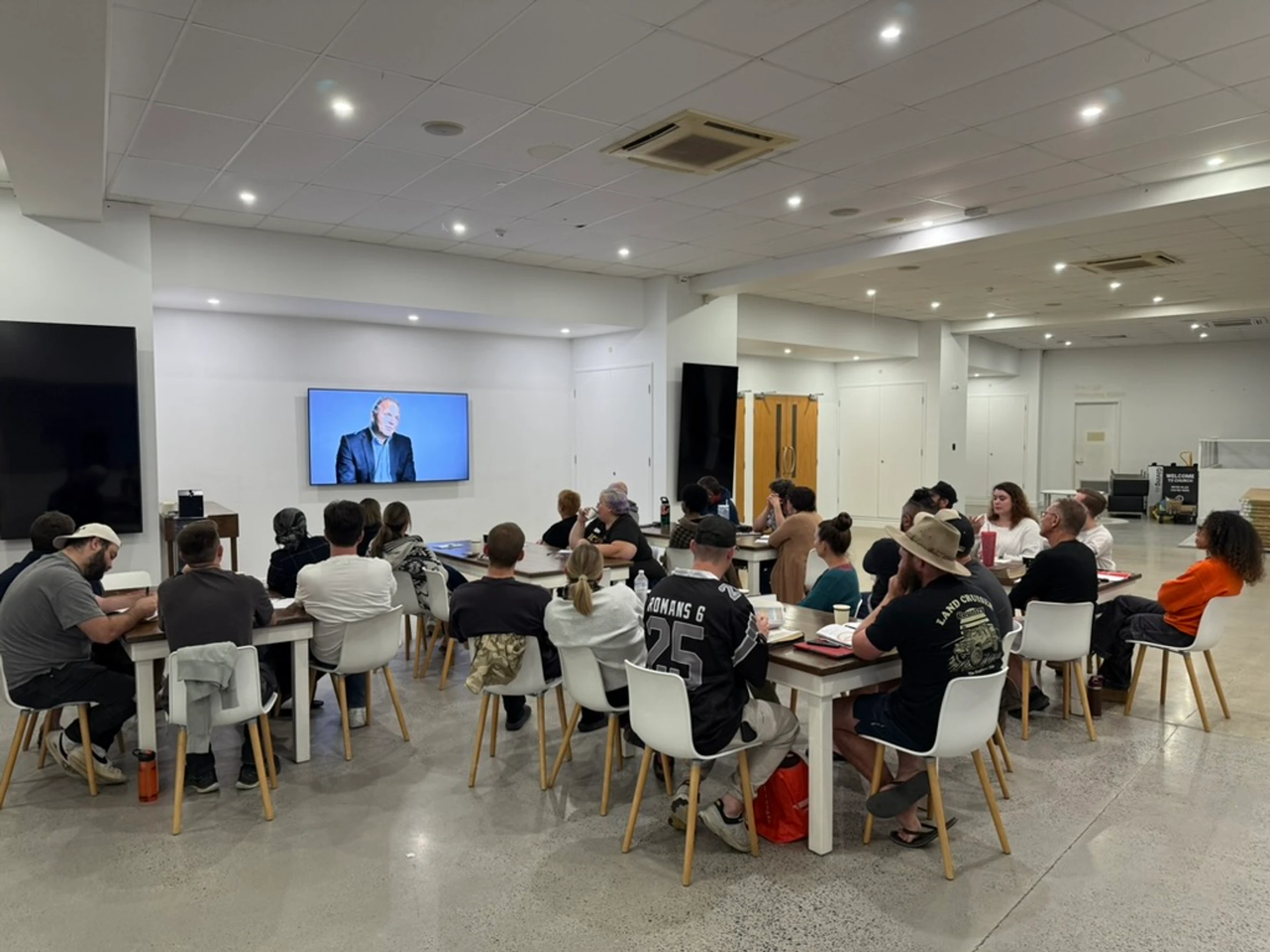 A diverse group of people seated at long tables in a well-lit room, watching a man give a presentation on a large wall-mounted screen.