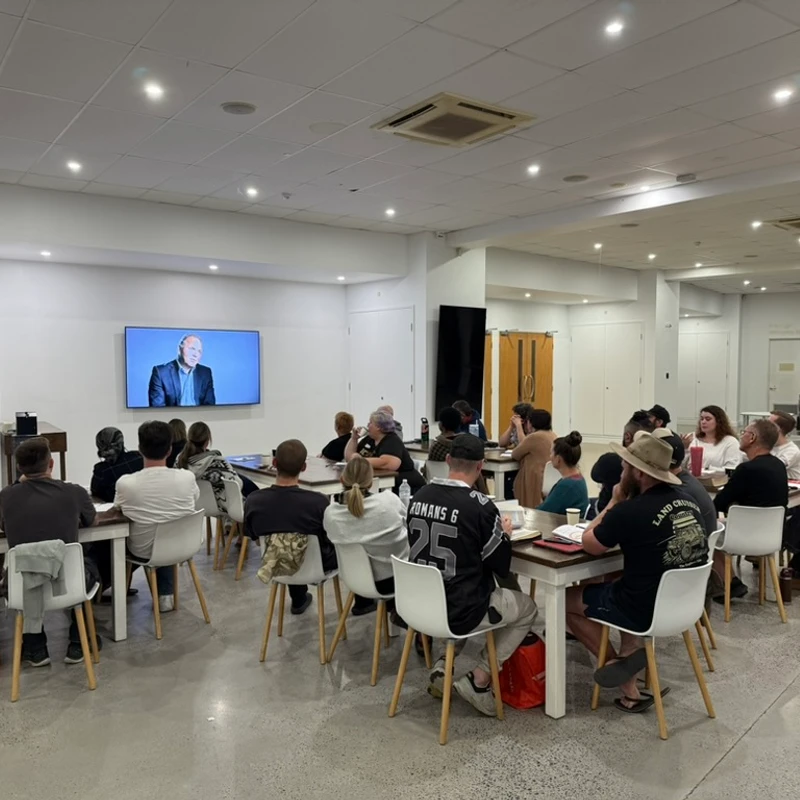 A diverse group of people seated at long tables in a well-lit room, watching a man give a presentation on a large wall-mounted screen.