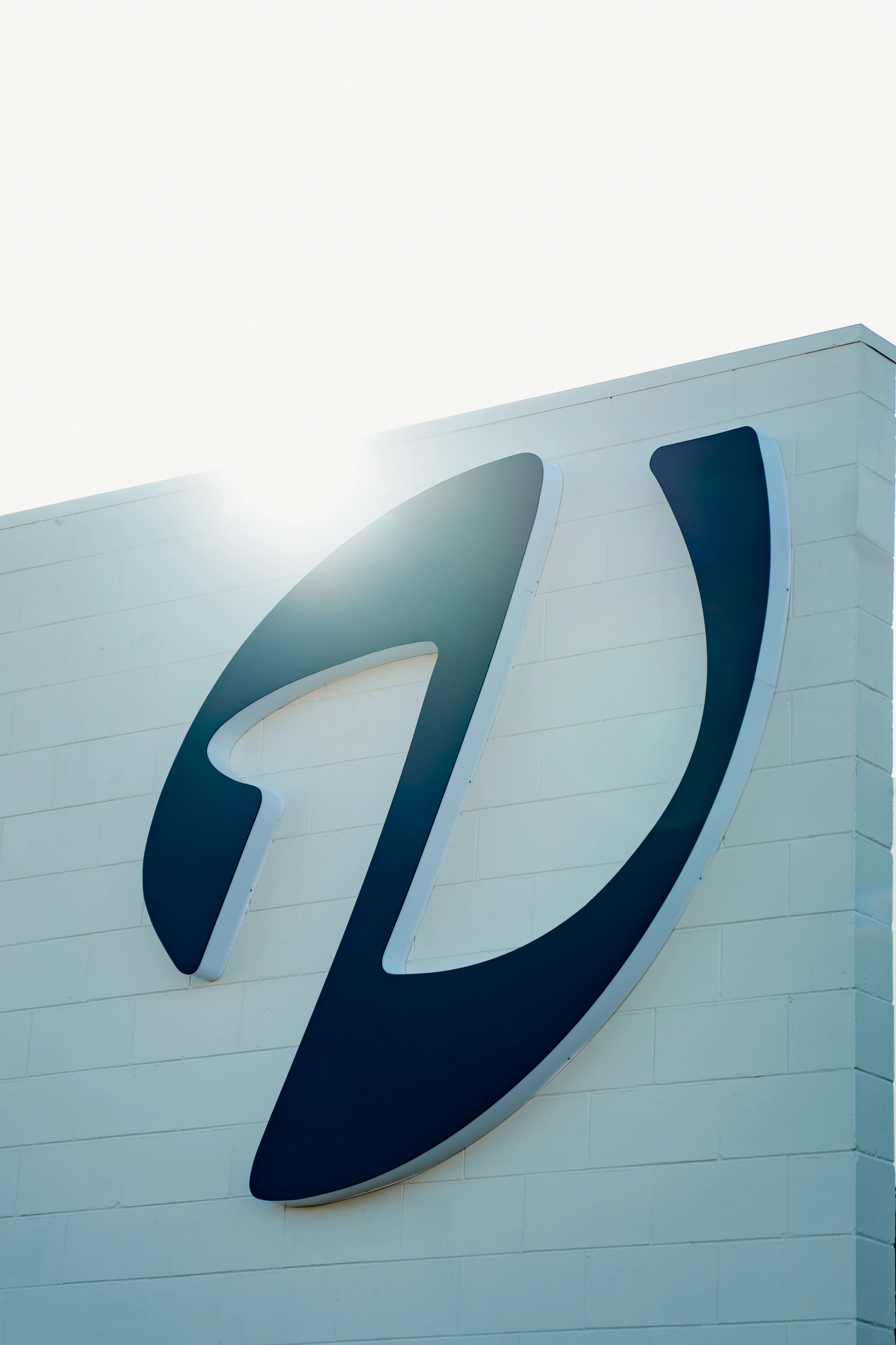 A low-angle shot of a large, abstract dark blue logo mounted on a white brick building wall. The sun flares brightly from behind the top of the logo.