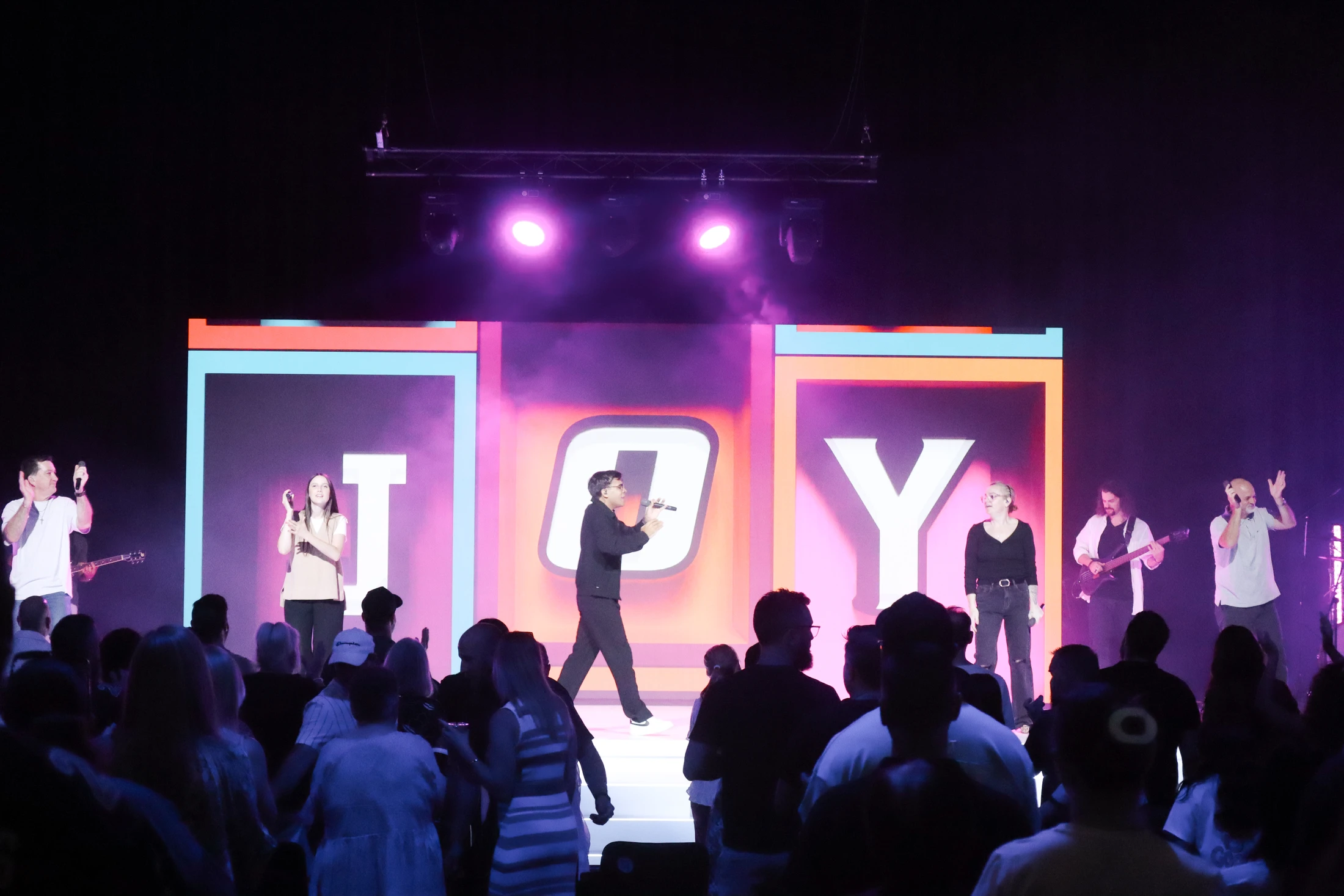 A band with several singers performs on a purple-lit stage for a crowd. A large screen behind them spells out the word 'JOY' in colorful block letters.