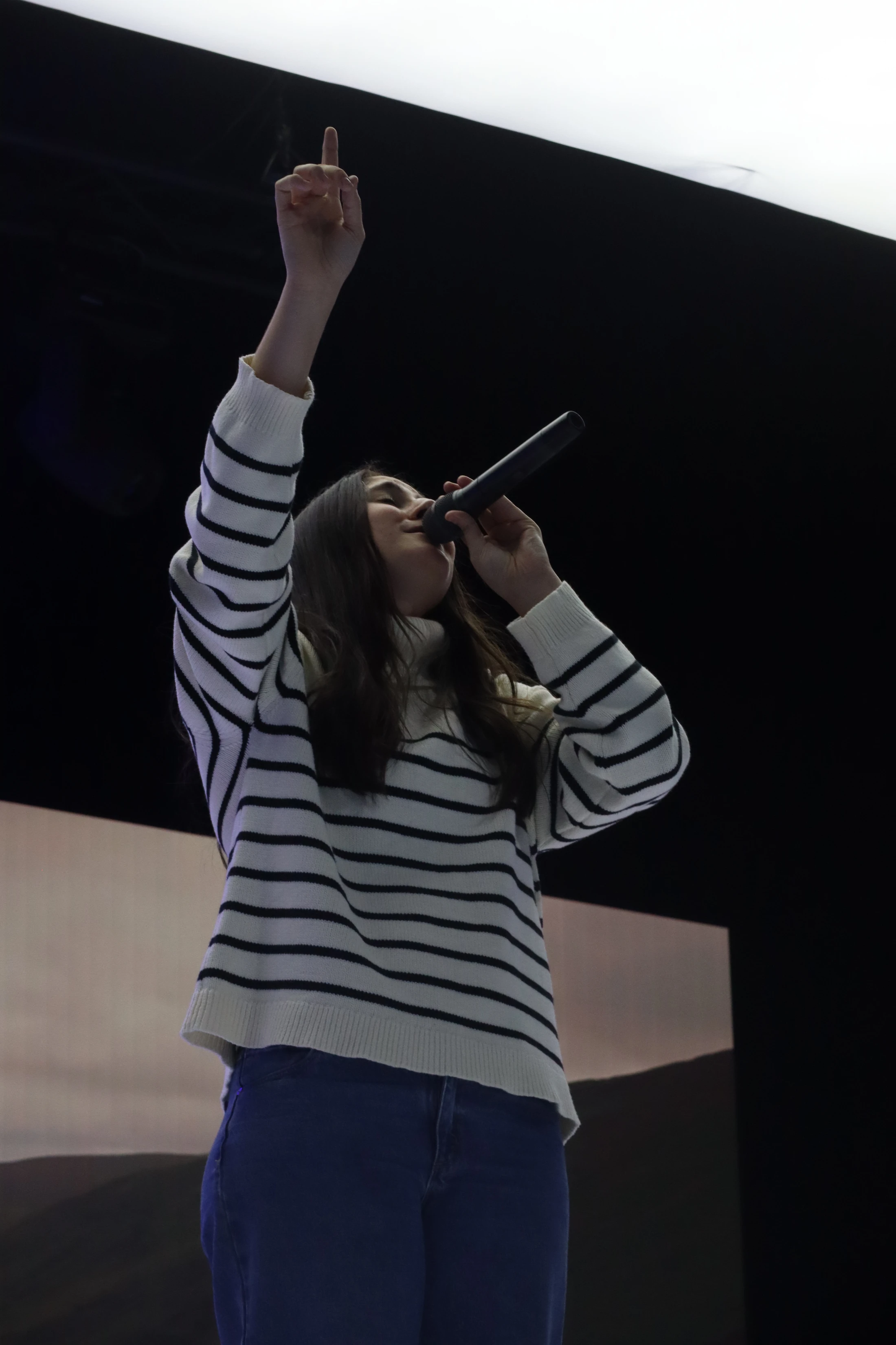 A young woman in a striped sweater and jeans sings into a microphone on a dimly lit stage. She raises one arm with her finger pointing upwards.