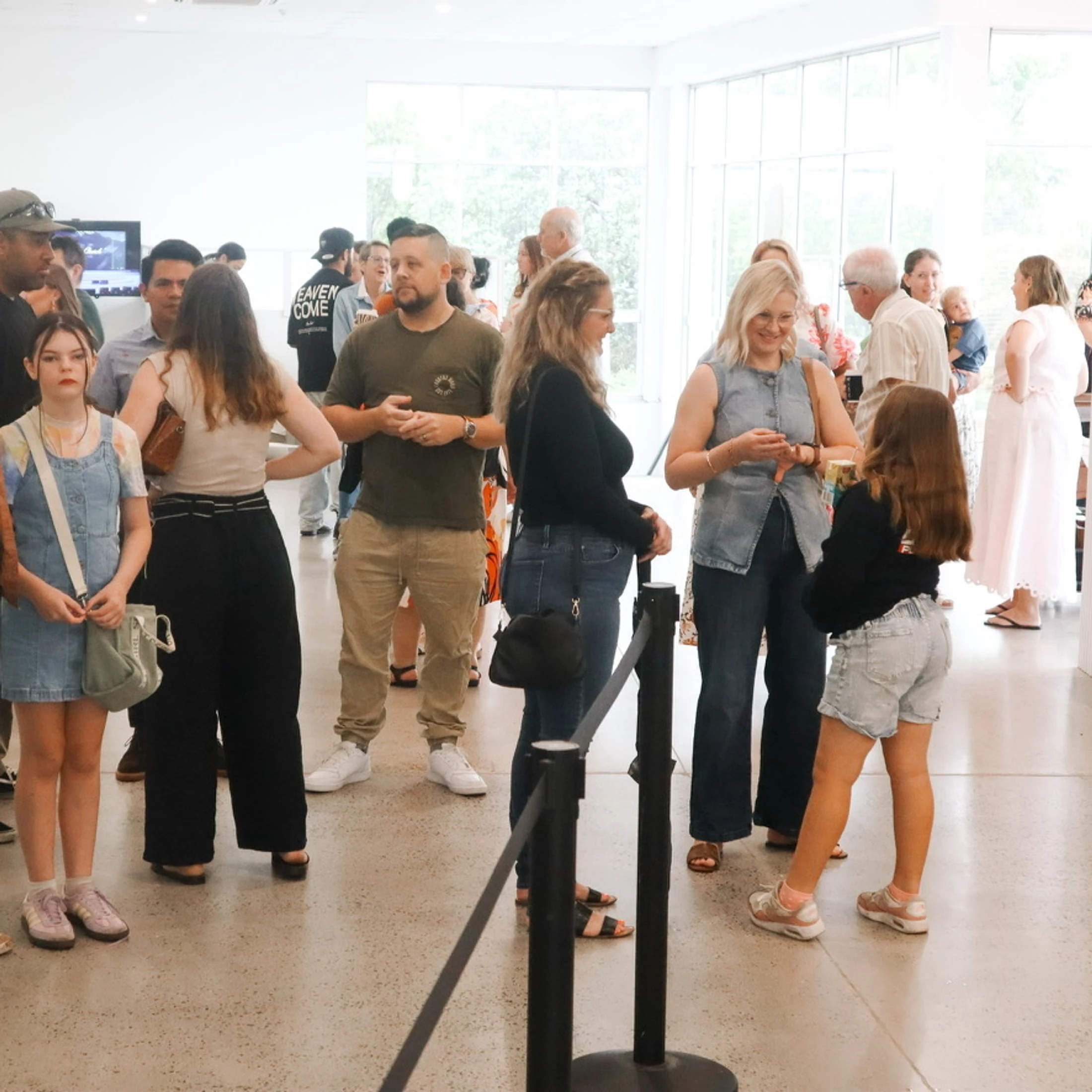 A diverse group of casually dressed people of all ages stand and talk in a bright, modern room with large windows and a polished concrete floor.