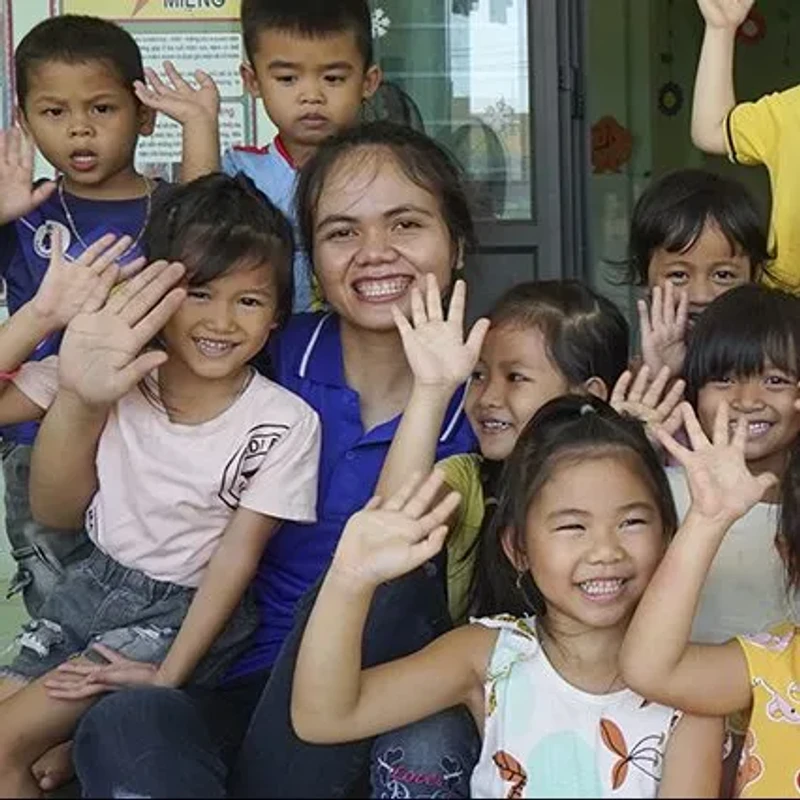 A smiling woman sits surrounded by a group of happy young children. They are all looking toward the camera and waving their hands in a friendly greeting.