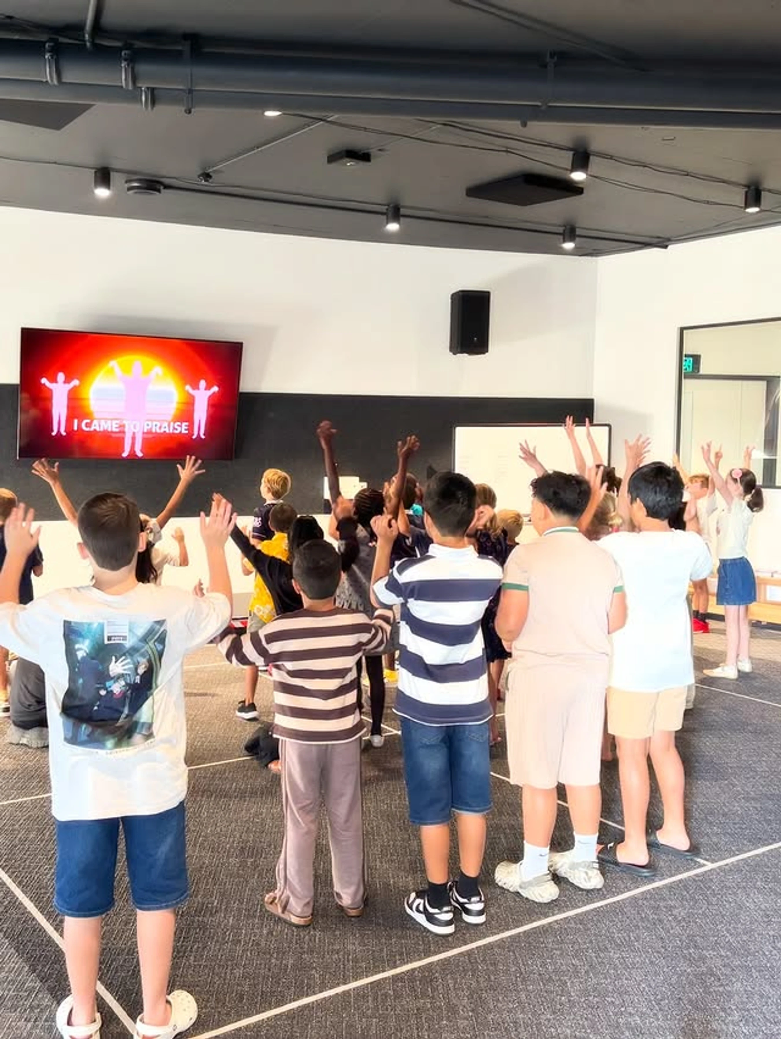 A group of children seen from behind, raising their hands in a room while facing a screen that says 'I CAME TO PRAISE'.
