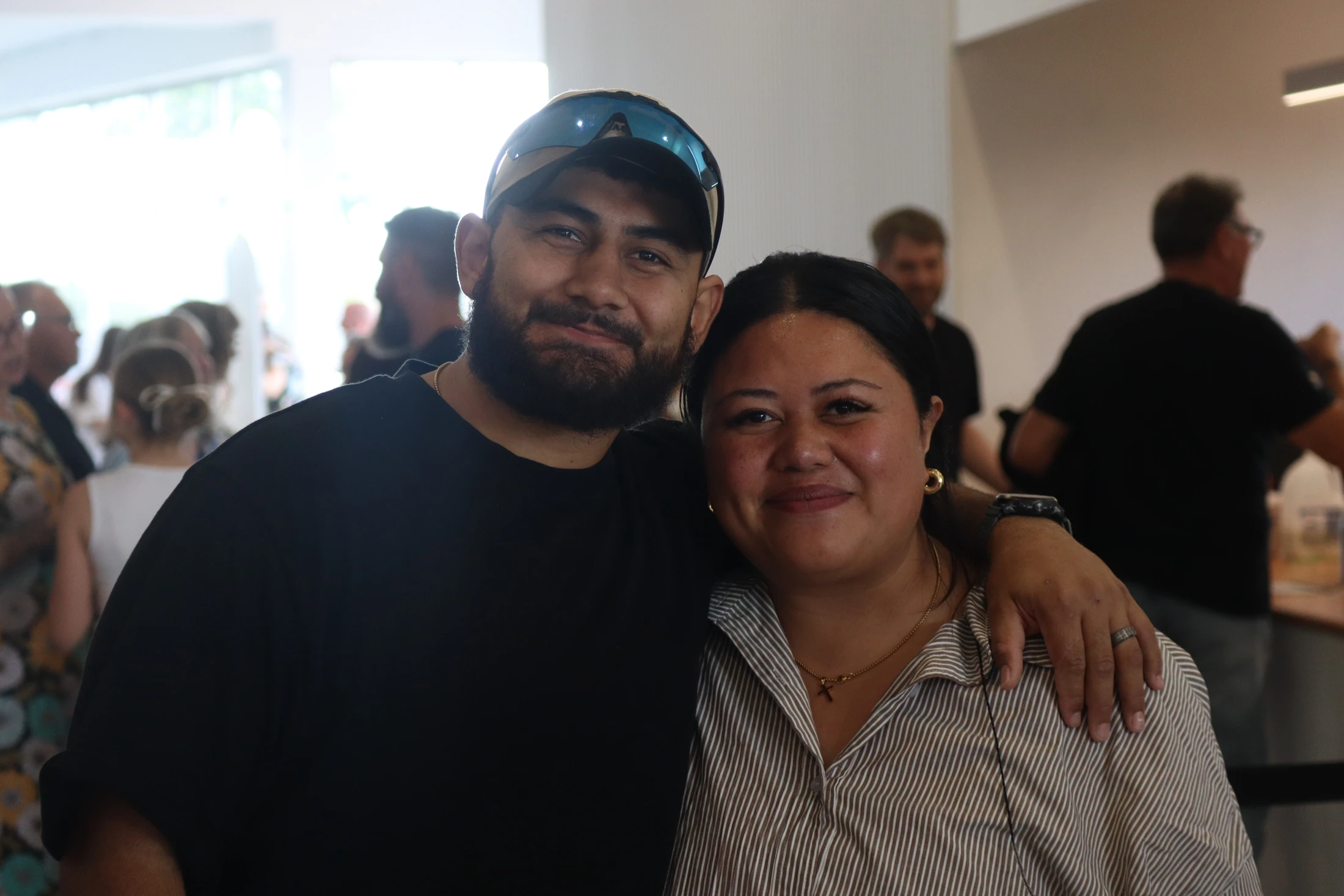 A smiling man with a beard and cap has his arm around a smiling woman in a striped shirt. They are posing for a photo indoors with people in the background.